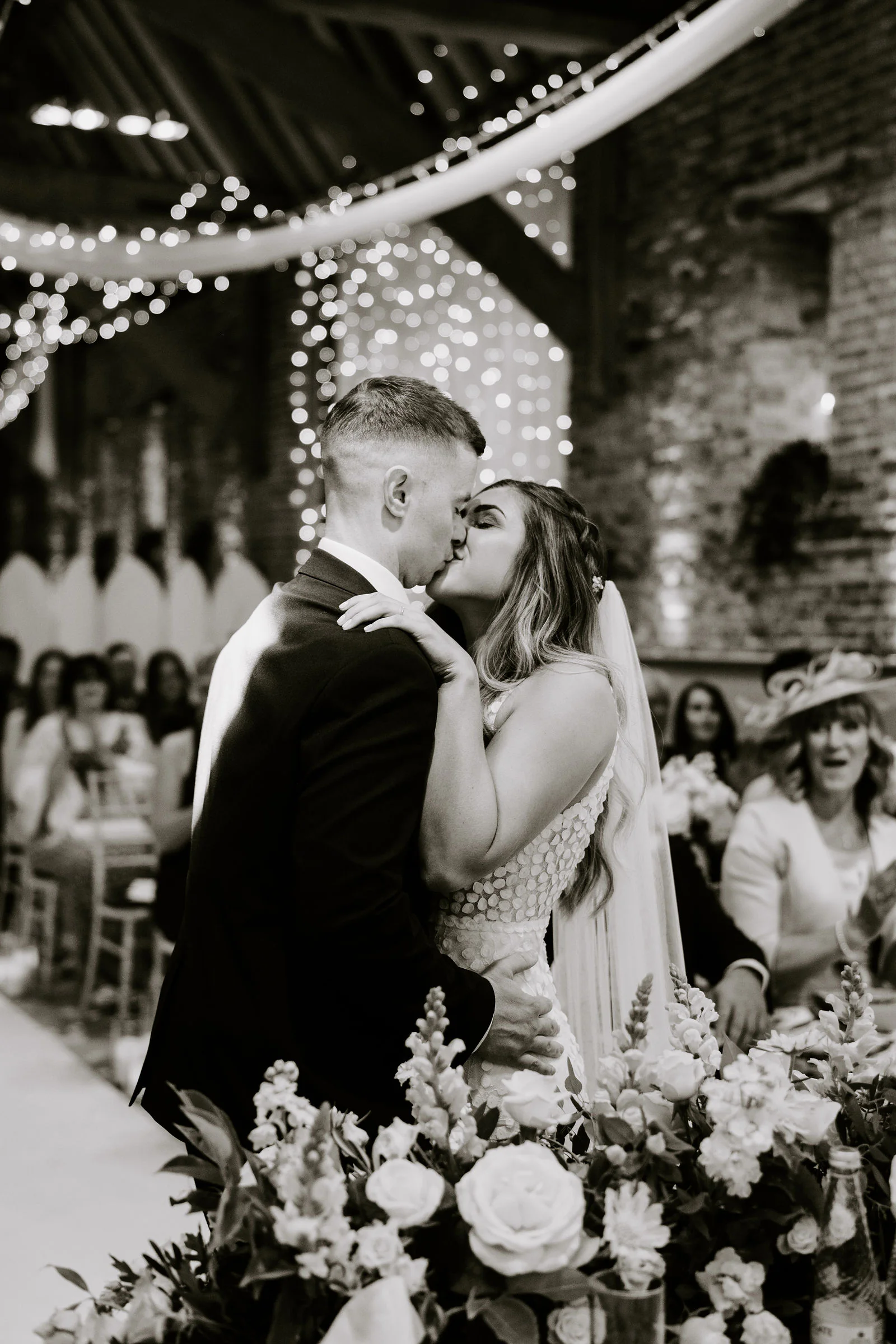 In black and white, Karl and Shantelle kiss during their ceremony beneath swags of fairy lights in a brick barn, guests in hats behind them