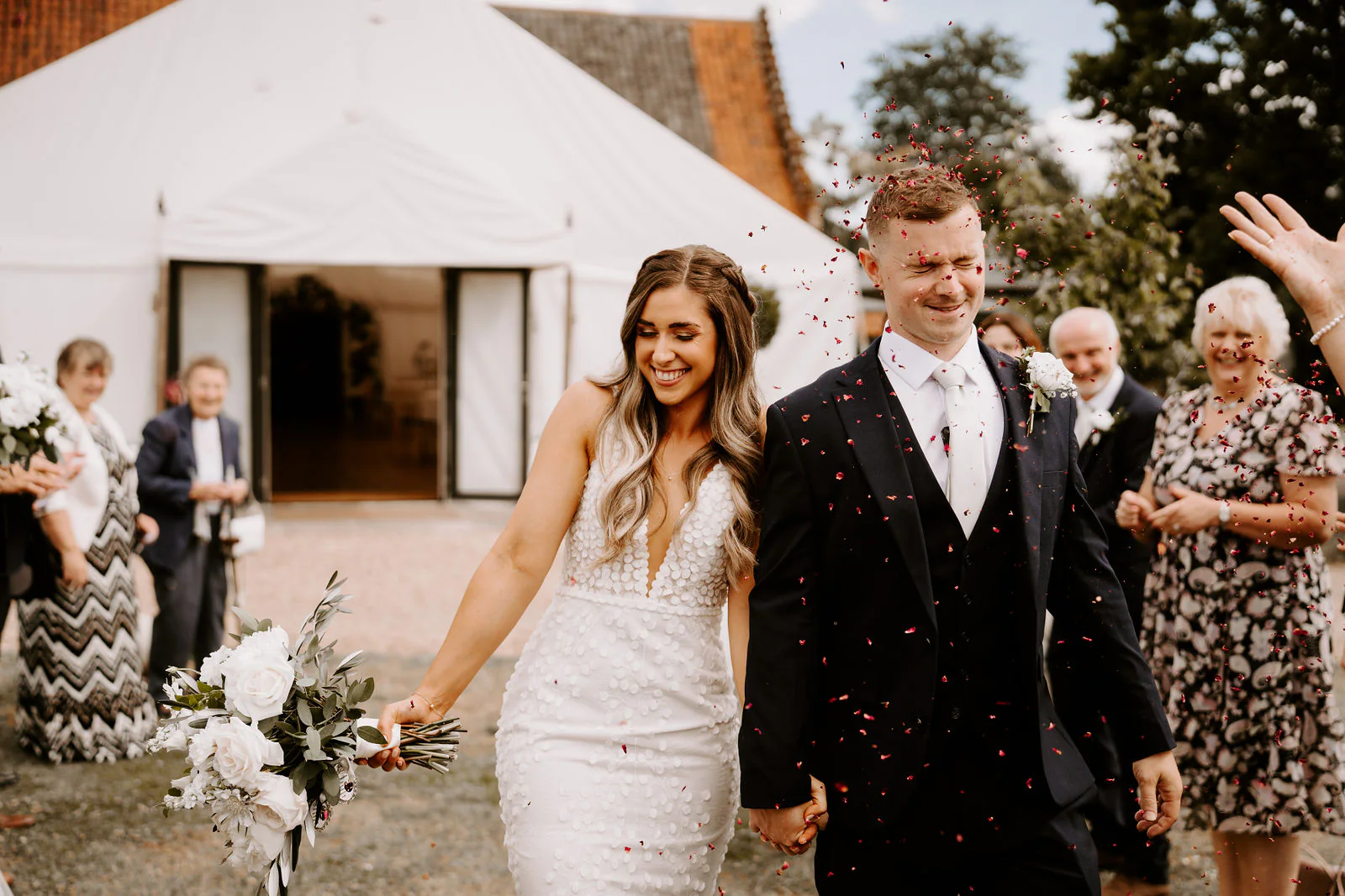 Shantelle and Karl walk hand in hand through confetti outside a white marquee as guests smile around them