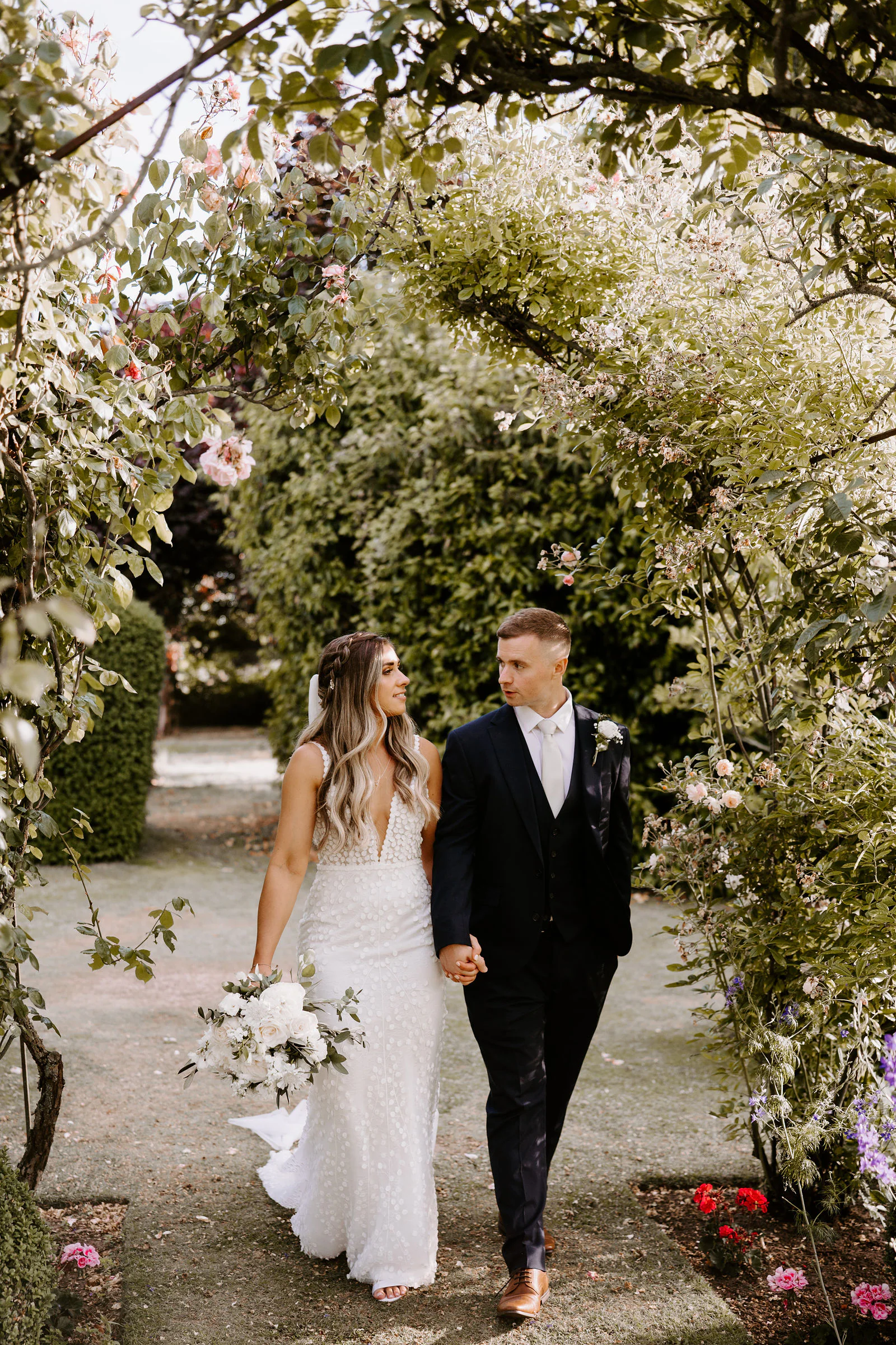 Shantelle and Karl walk hand in hand beneath an arch of roses and greenery along a gravel path