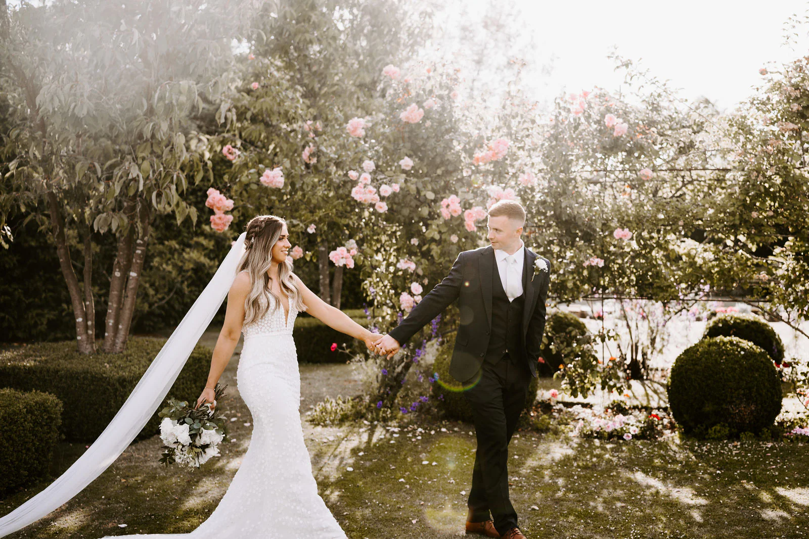 Shantelle leads Karl by the hand through a garden of pink roses and clipped box, her long veil trailing behind her