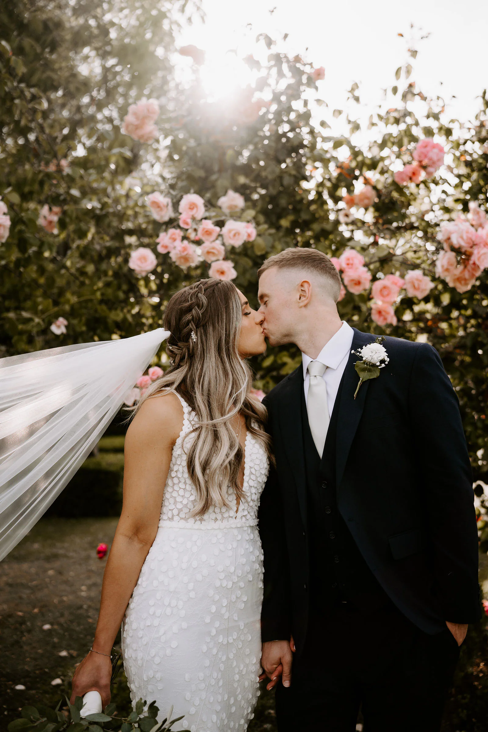 Shantelle and Karl kiss in front of pink roses with the sun behind them, her veil lifting at the edge of the frame