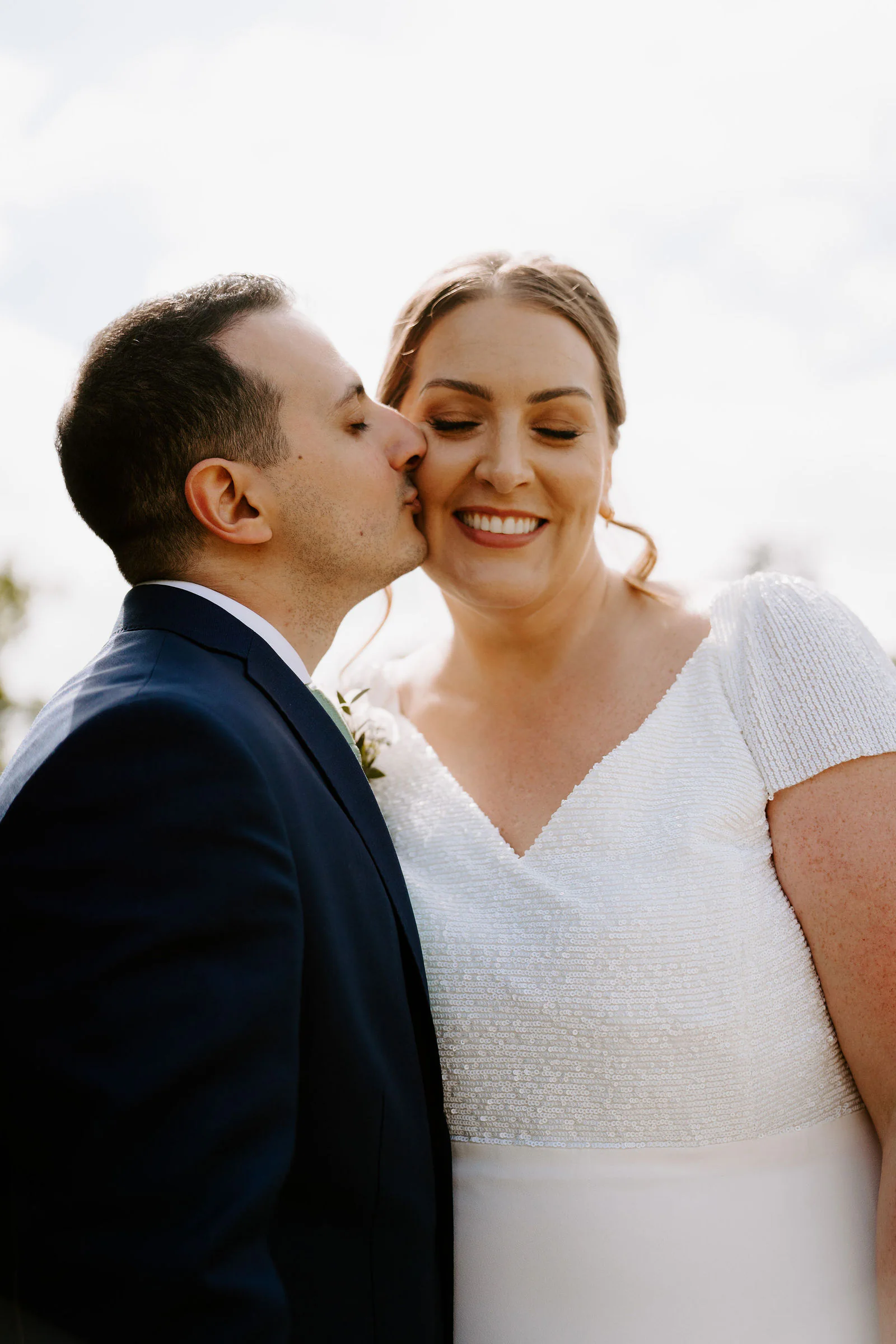 Thierry in a navy suit kisses Aimee on the cheek as she smiles with her eyes closed in soft light at Hengrave Hall