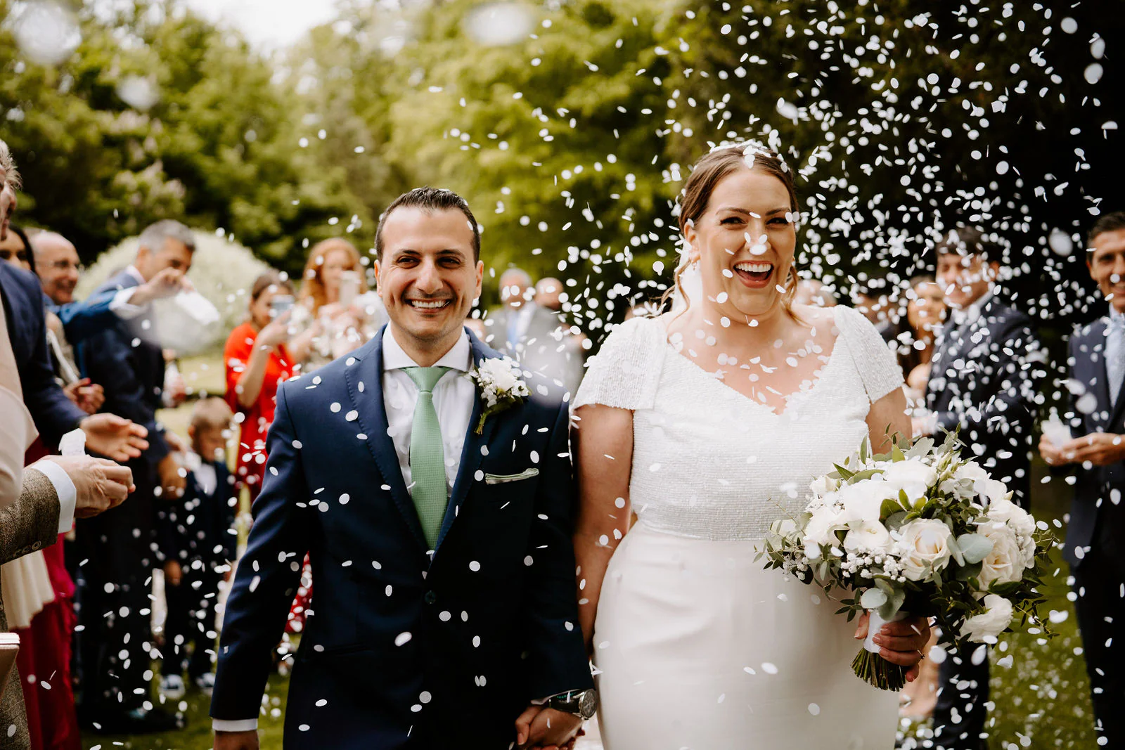 Aimee and Thierry laugh as they walk hand in hand through a cloud of white confetti between rows of guests on the lawn at Hengrave Hall