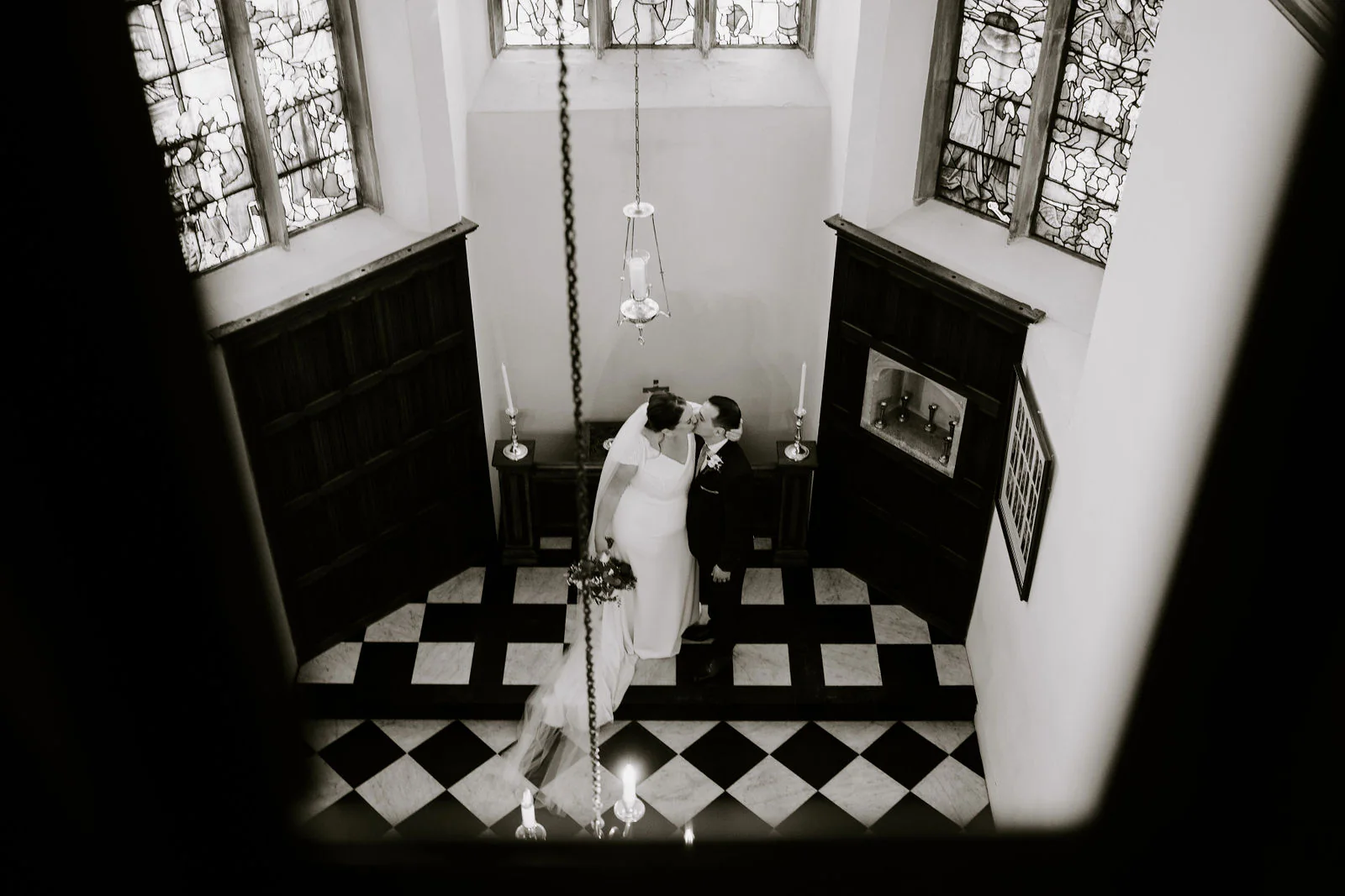 In black and white, seen from a landing above, Aimee and Thierry kiss on a chequered floor beneath leaded windows at Hengrave Hall