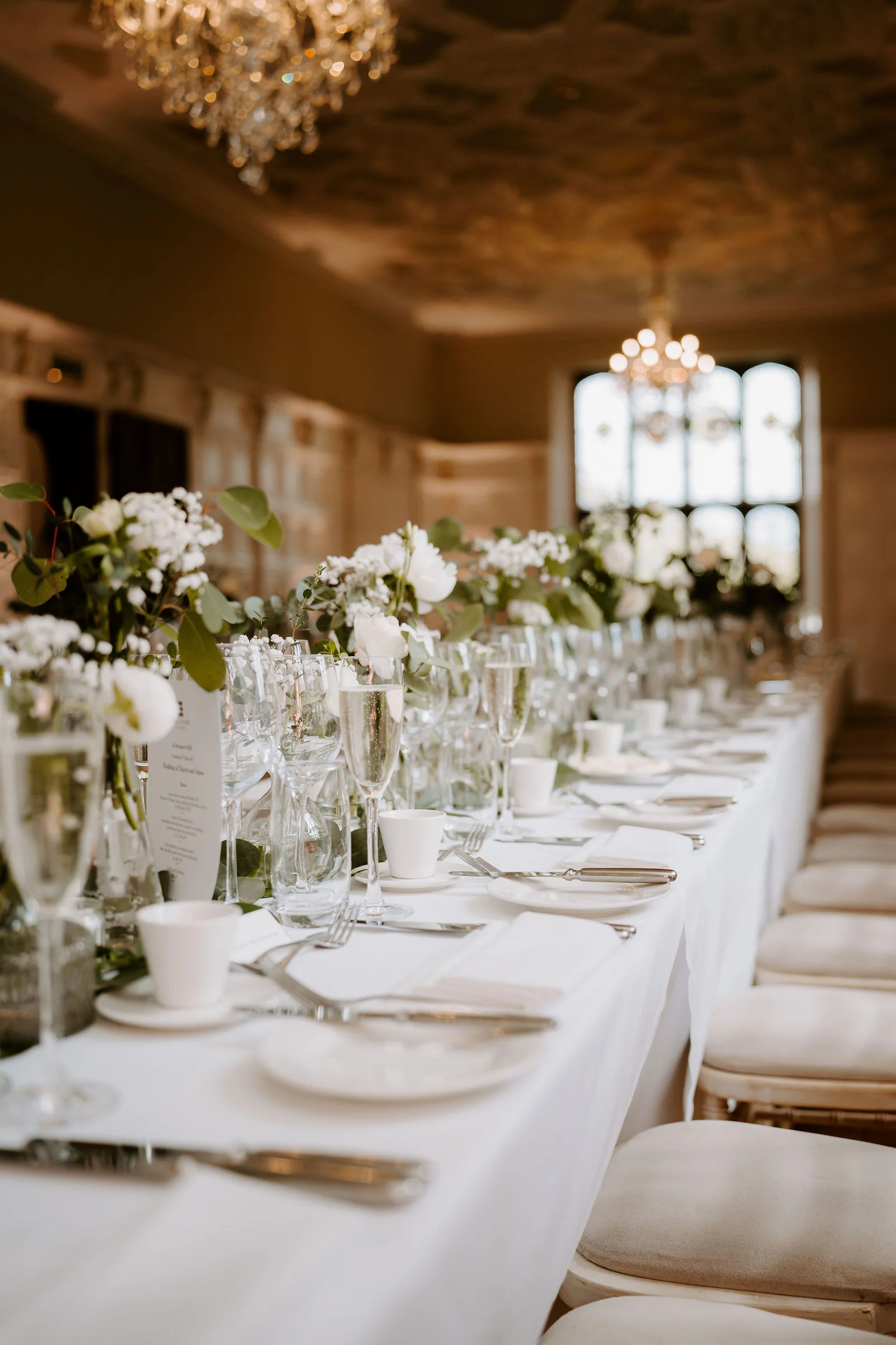 A long table laid with white linen, glasses and flowers beneath a chandelier in a panelled room at Hengrave Hall