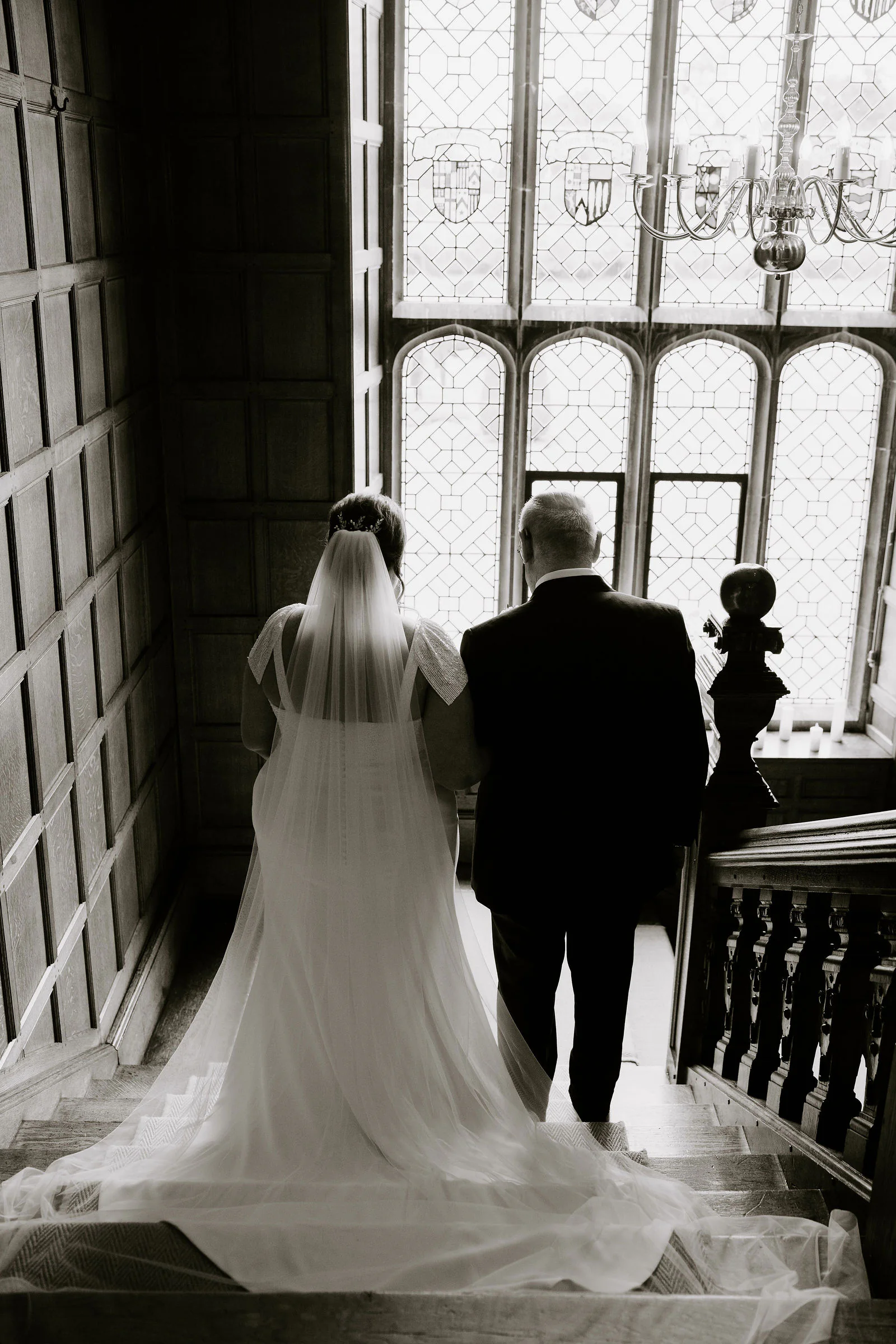 In black and white, Aimee in a long veil and train descends a dark panelled staircase beside a man in a dark suit towards a tall leaded window