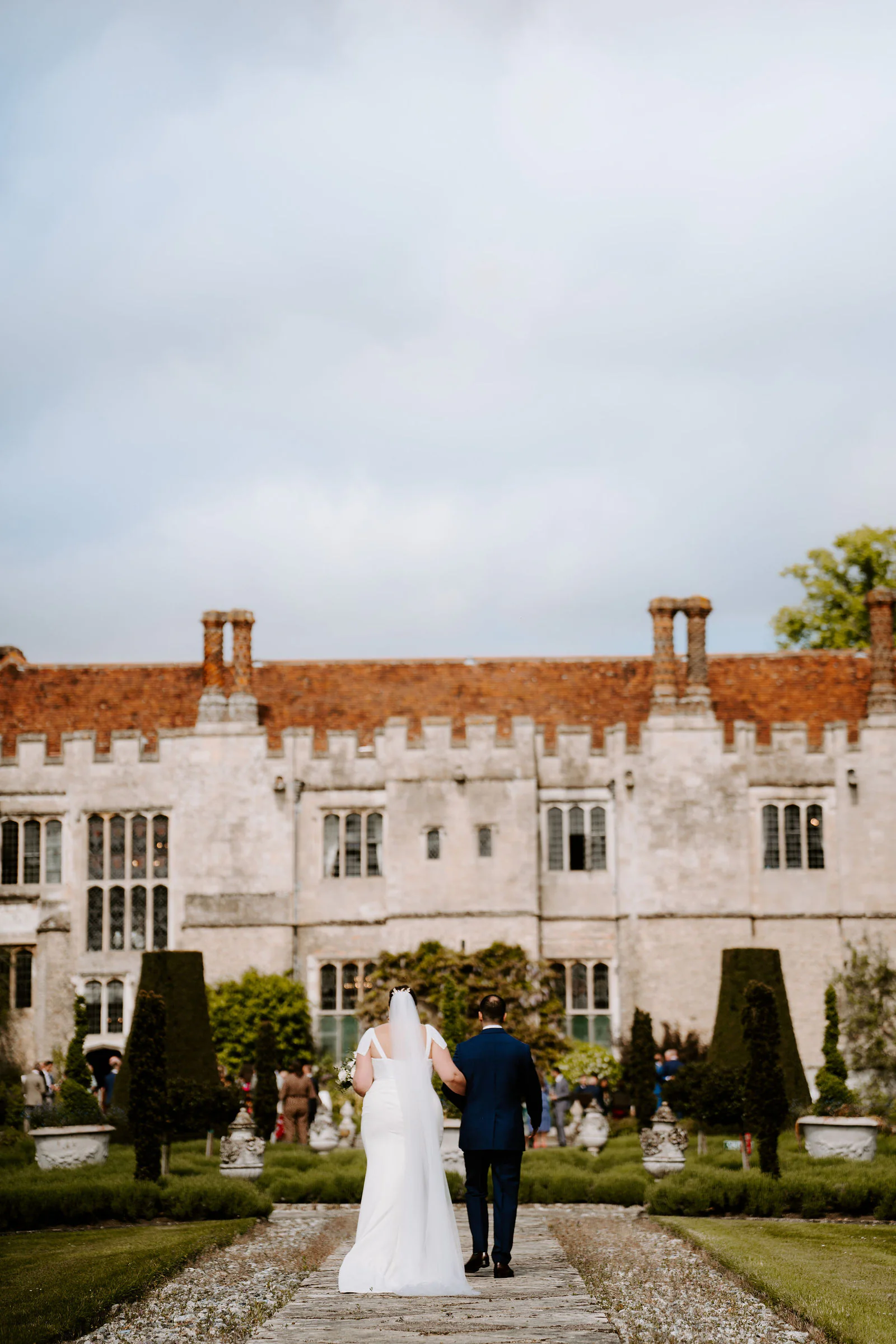 Aimee and Thierry walk away from the camera along a gravel path through formal clipped gardens towards the red-brick and stone front of Hengrave Hall