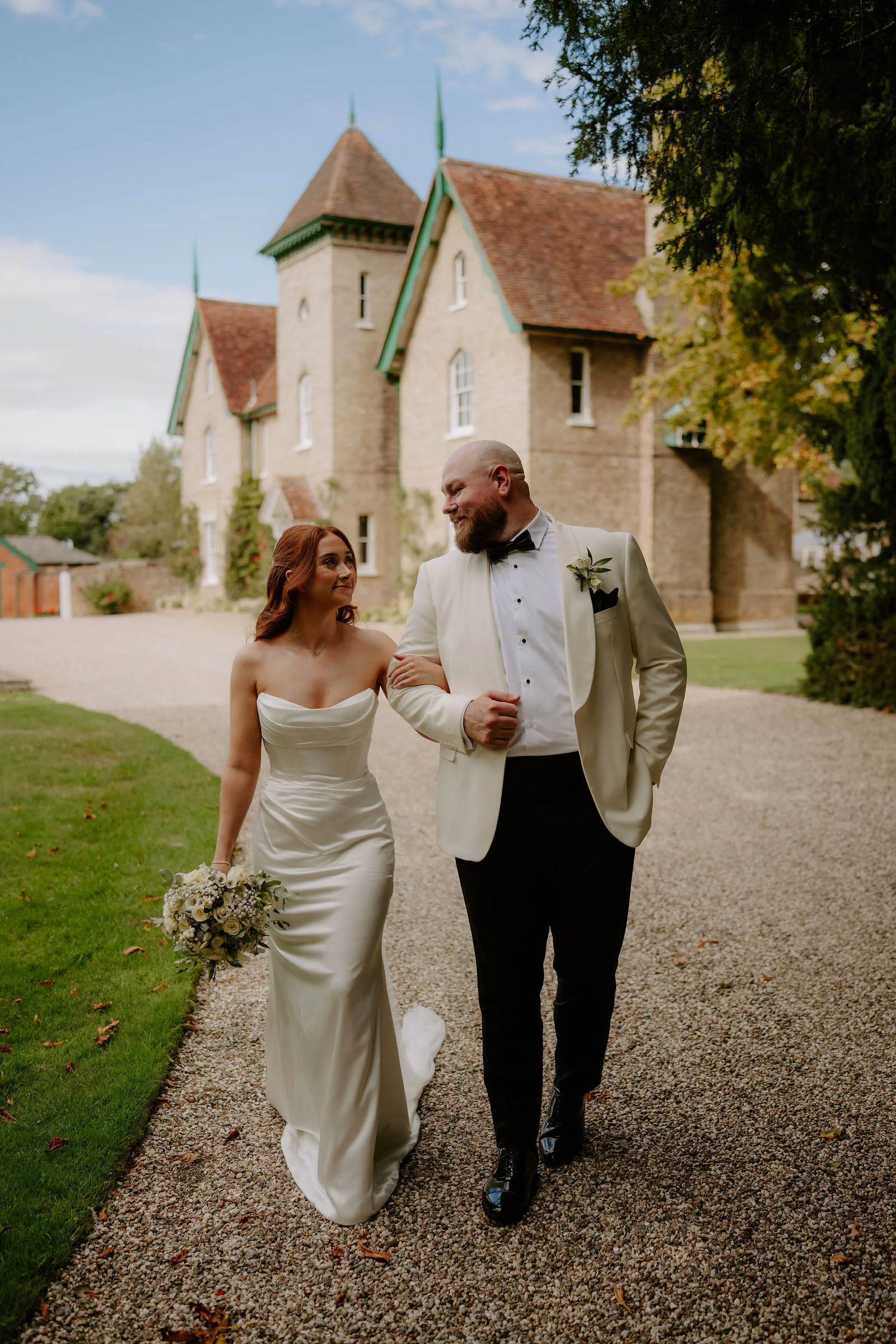 Katie and Mitch walk arm in arm along a gravel drive with the stone house and tower at Smeetham Hall behind them, Katie holding a bouquet of white flowers