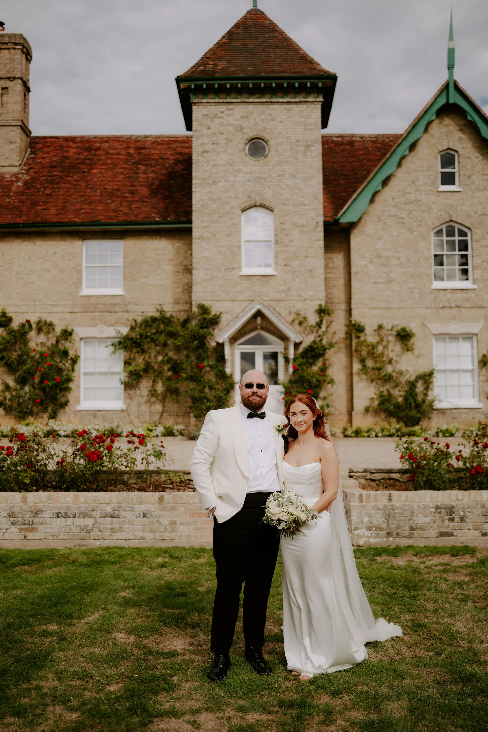 Katie and Mitch stand close together on the lawn in front of the stone house at Smeetham Hall, with its tower, green-trimmed gables and climbing plants behind them