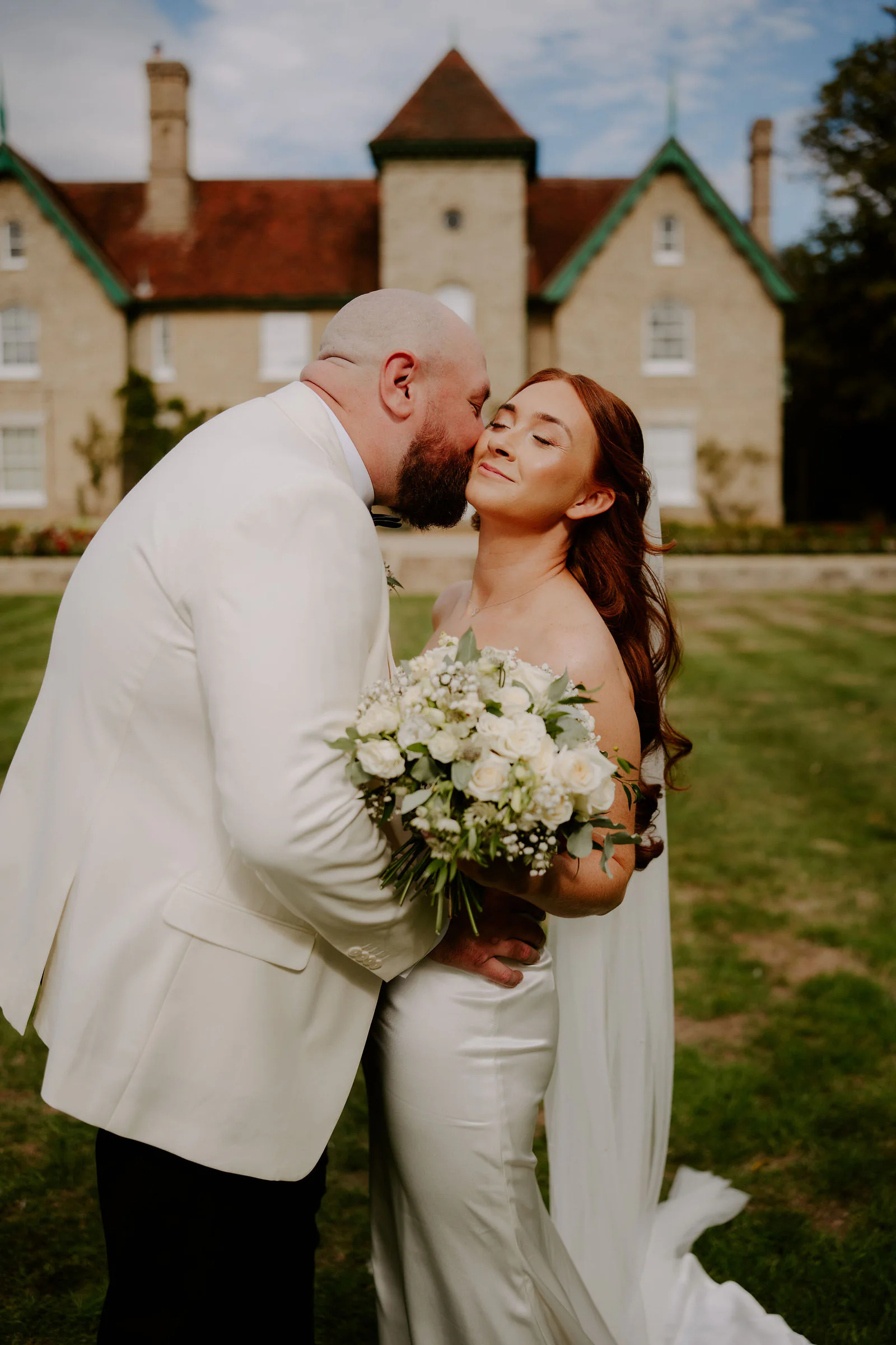 Mitch in a white dinner jacket kisses Katie on the cheek as she holds a bouquet of white flowers, with the stone house at Smeetham Hall and its tower behind them