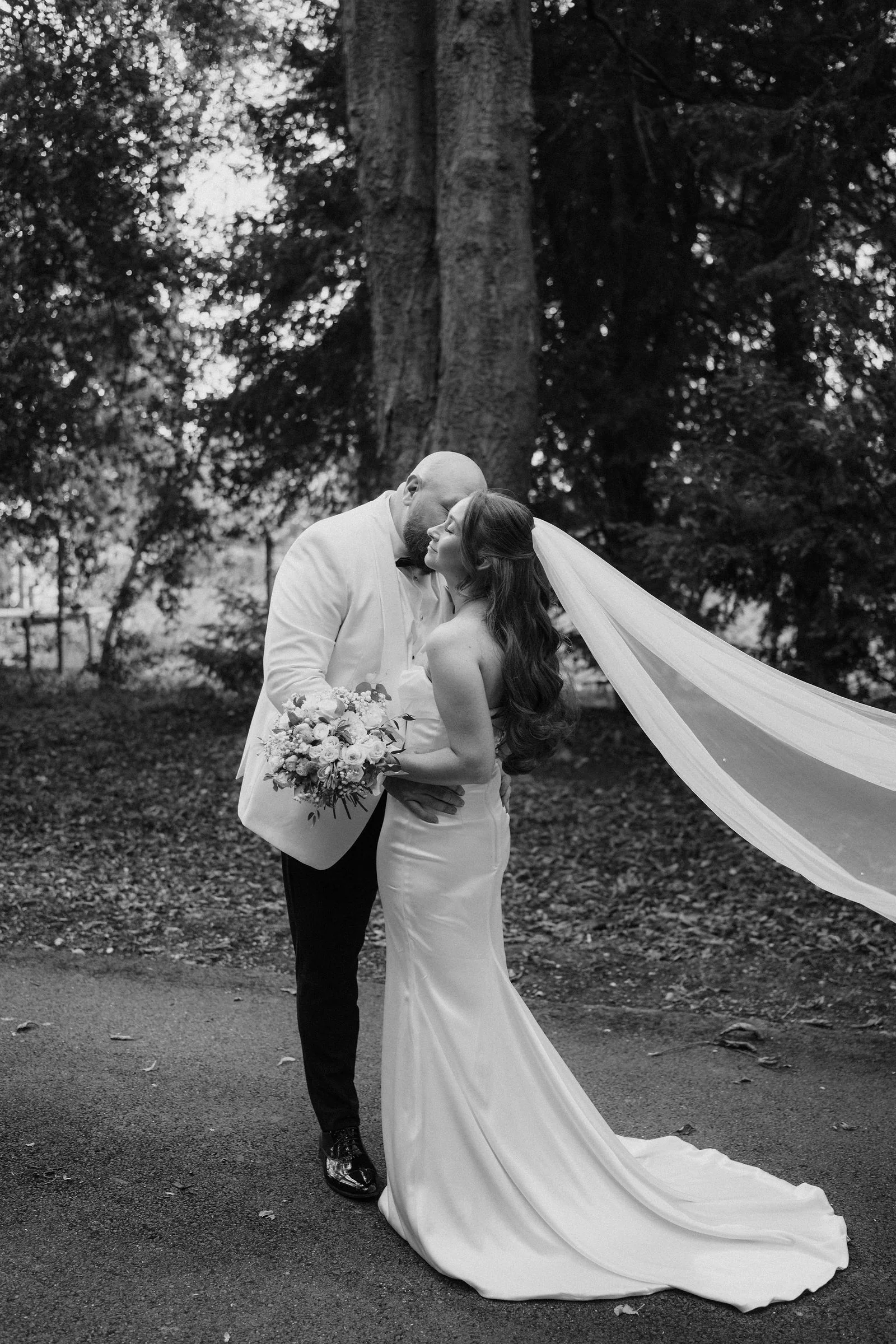 In black and white, Mitch kisses Katie beneath tall trees as her long veil streams out beside them