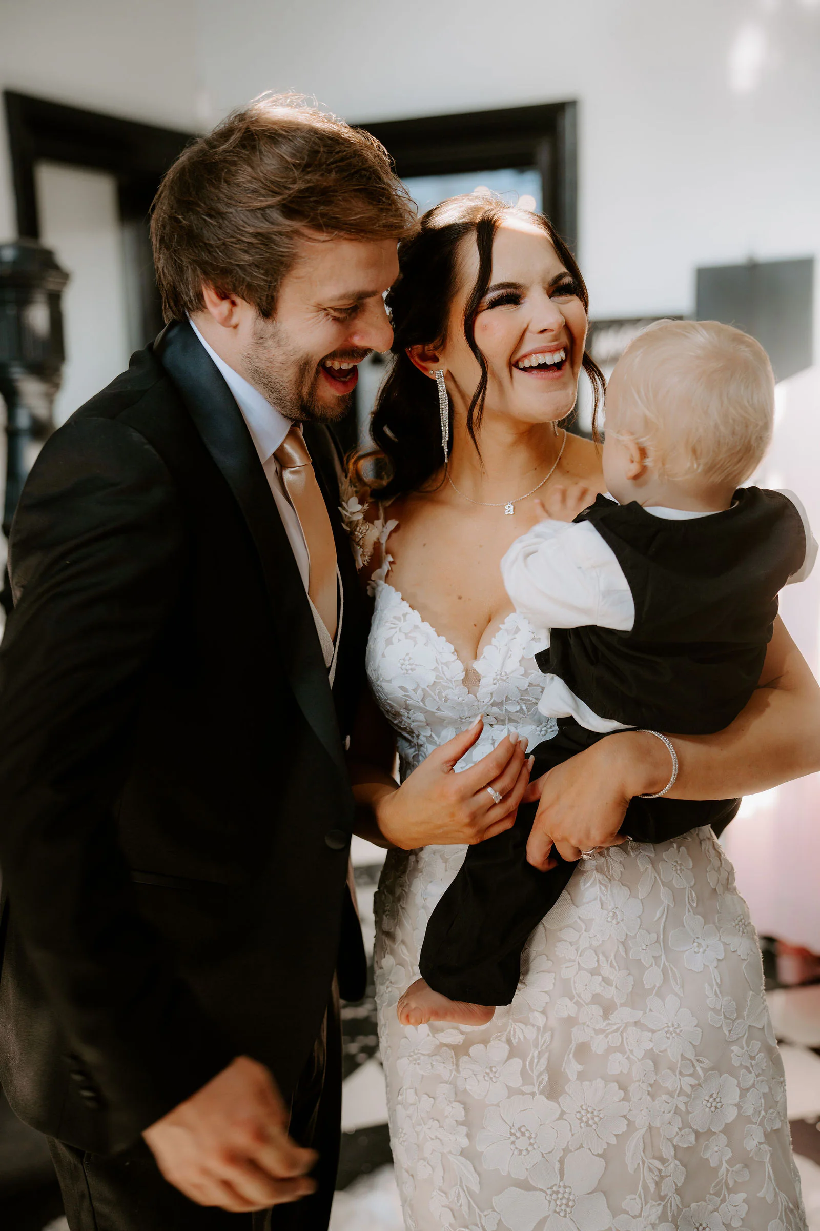 Anthony and Paige laugh together as Paige holds a baby in a black outfit inside the manor