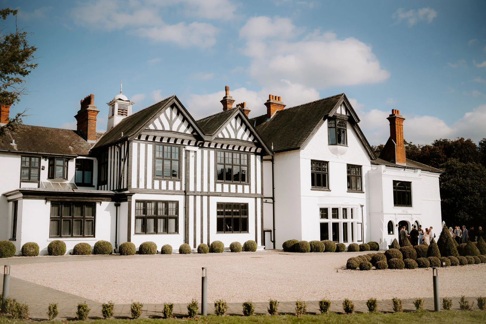 The black and white timbered front of Swynford Manor with its gabled roofs and tall chimneys, seen across a gravel drive under a bright blue sky
