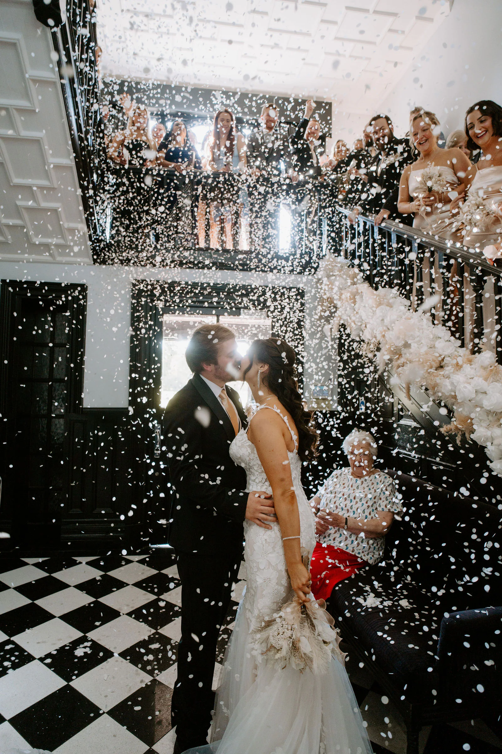 Anthony and Paige kiss on a black and white chequered floor beneath a staircase as guests on the landing above throw clouds of confetti