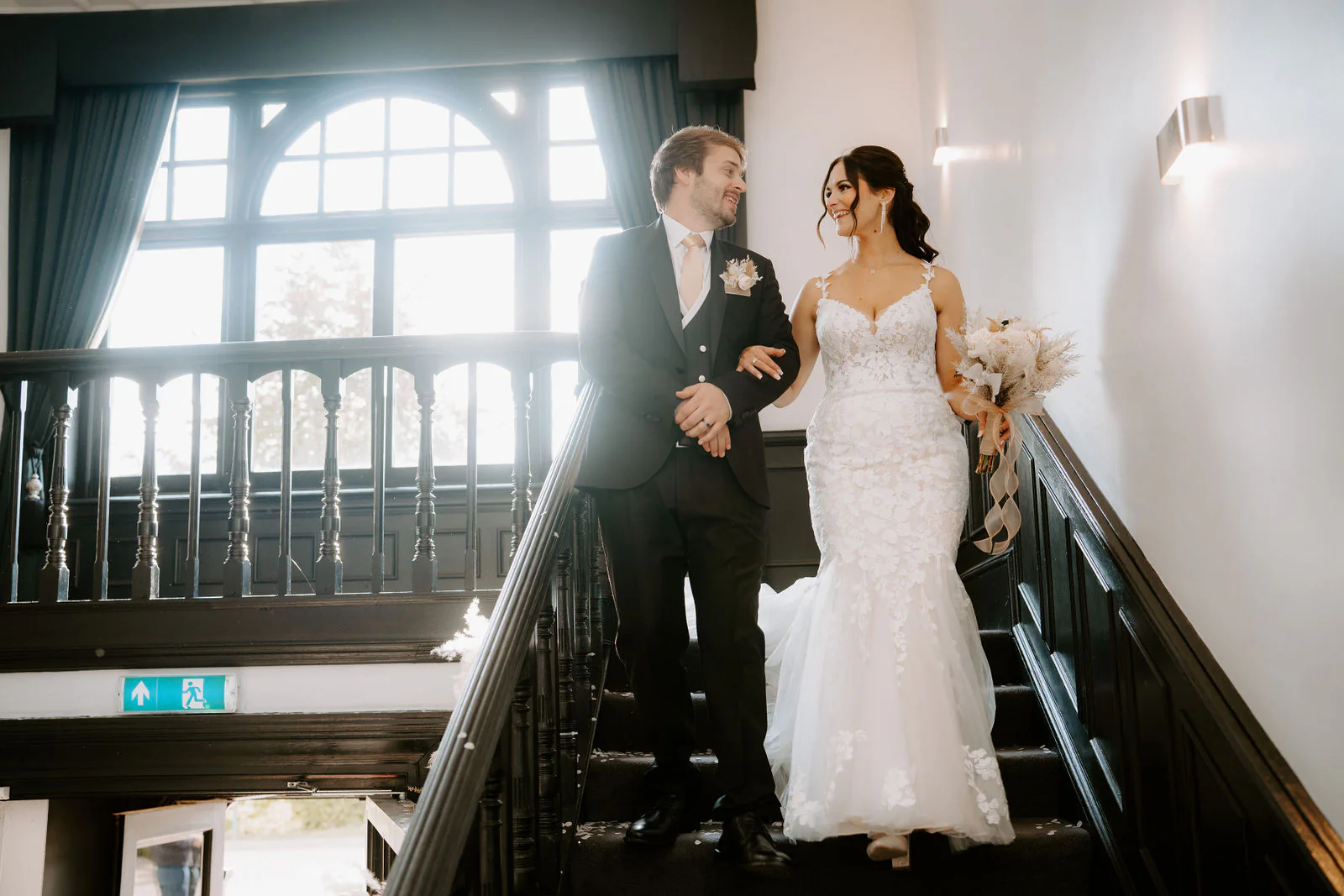 Anthony and Paige walk arm in arm down a dark wooden staircase beside a large arched window