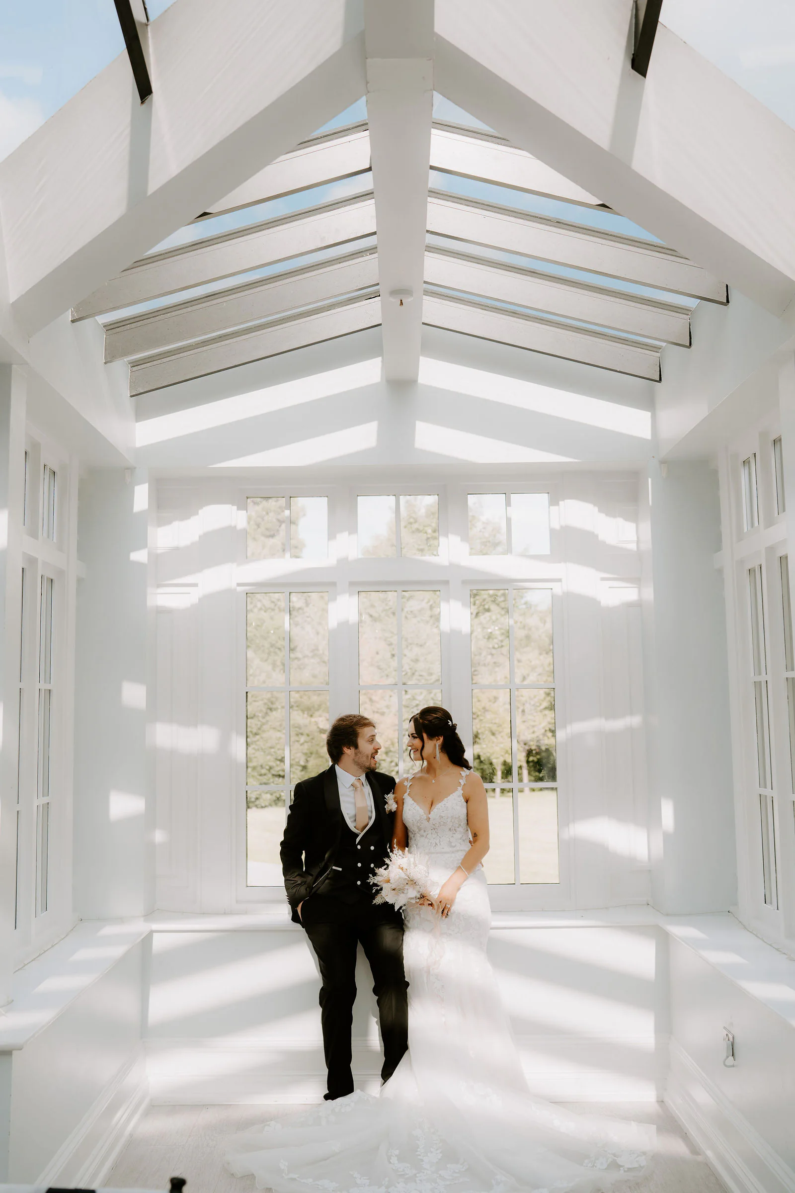 Anthony and Paige stand together in a bright white glazed room with a pitched glass roof and tall windows, light falling in stripes across the floor