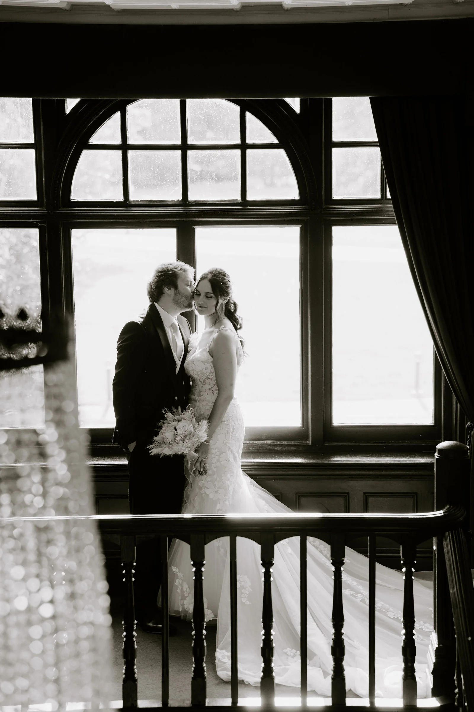 In black and white, Anthony and Paige embrace in front of a large arched window above a dark banister