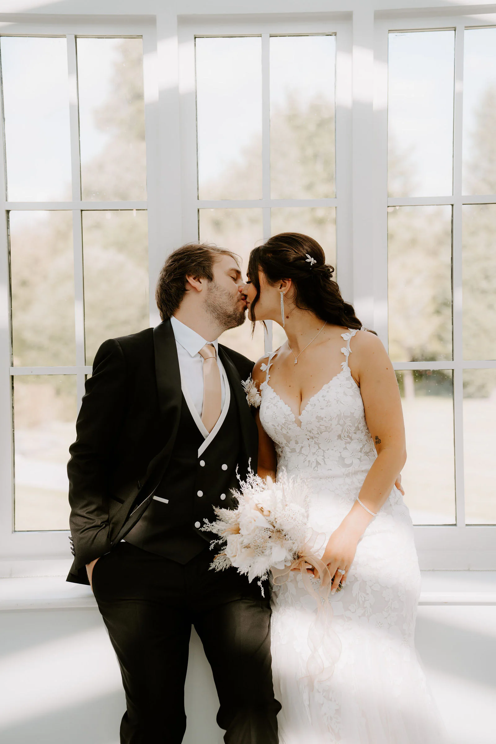 Anthony and Paige kiss in front of tall white-framed windows, Paige holding a bouquet of dried grasses and pale flowers