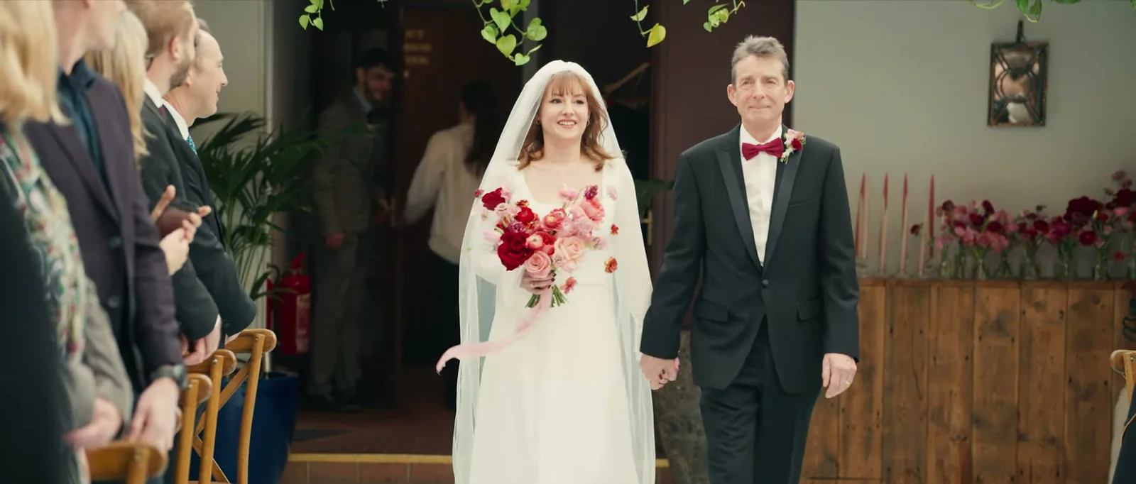 A smiling bride in a veil holding a pink and red bouquet walks hand in hand with a man in a red bow tie between rows of guests