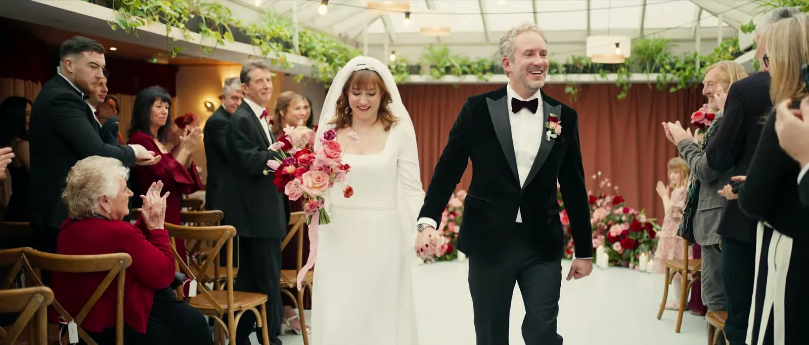 A groom and a bride in a veil walk hand in hand along the aisle, smiling, as guests on both sides applaud