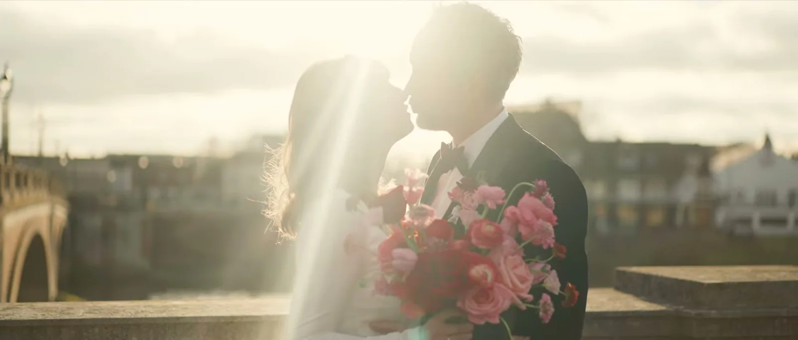 A bride and groom kiss in silhouette against the low winter sun on a bridge, the bride holding a pink and red bouquet