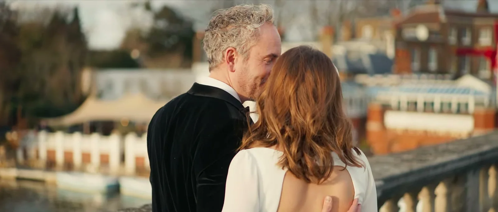 A groom kisses his bride's cheek on a stone bridge, with a riverside terrace and canopy softly out of focus behind them