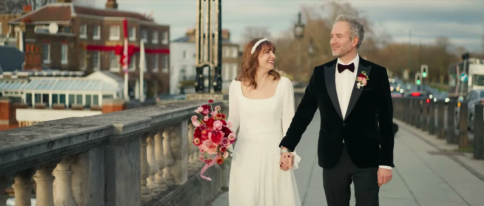A bride in a long white dress with a pale headband and a groom in a black velvet jacket and bow tie walk hand in hand along a stone bridge, smiling at each other, with riverside buildings behind them