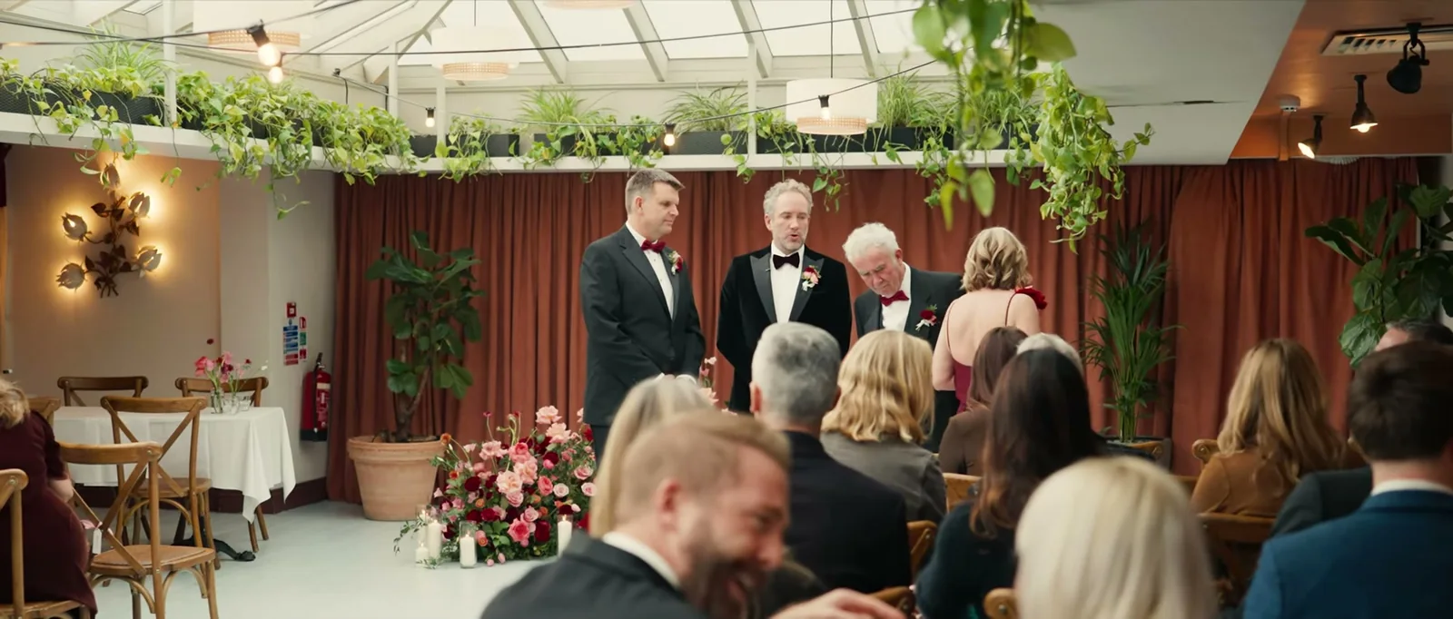 Three men in dark suits stand at the front of a glass-roofed room hung with trailing plants, in front of rust-coloured curtains and a mound of pink and red roses, as seated guests look on