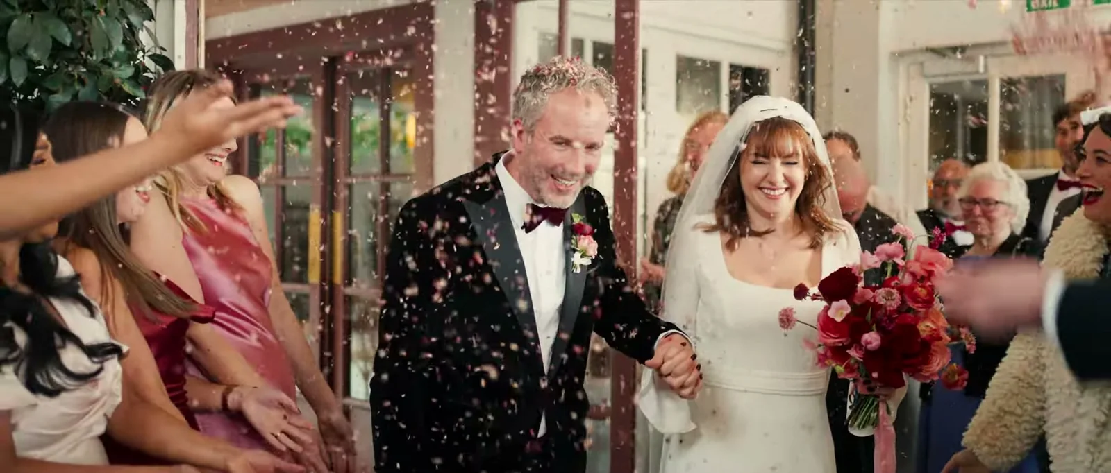 A laughing groom and bride in a veil walk through a shower of confetti, with guests around them and a bouquet of red and pink flowers