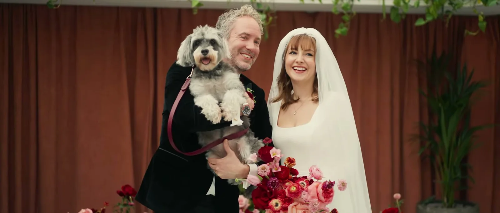 A groom holds a small grey dog in his arms beside his smiling bride in a veil, in front of rust-coloured curtains and a bouquet of red and pink flowers