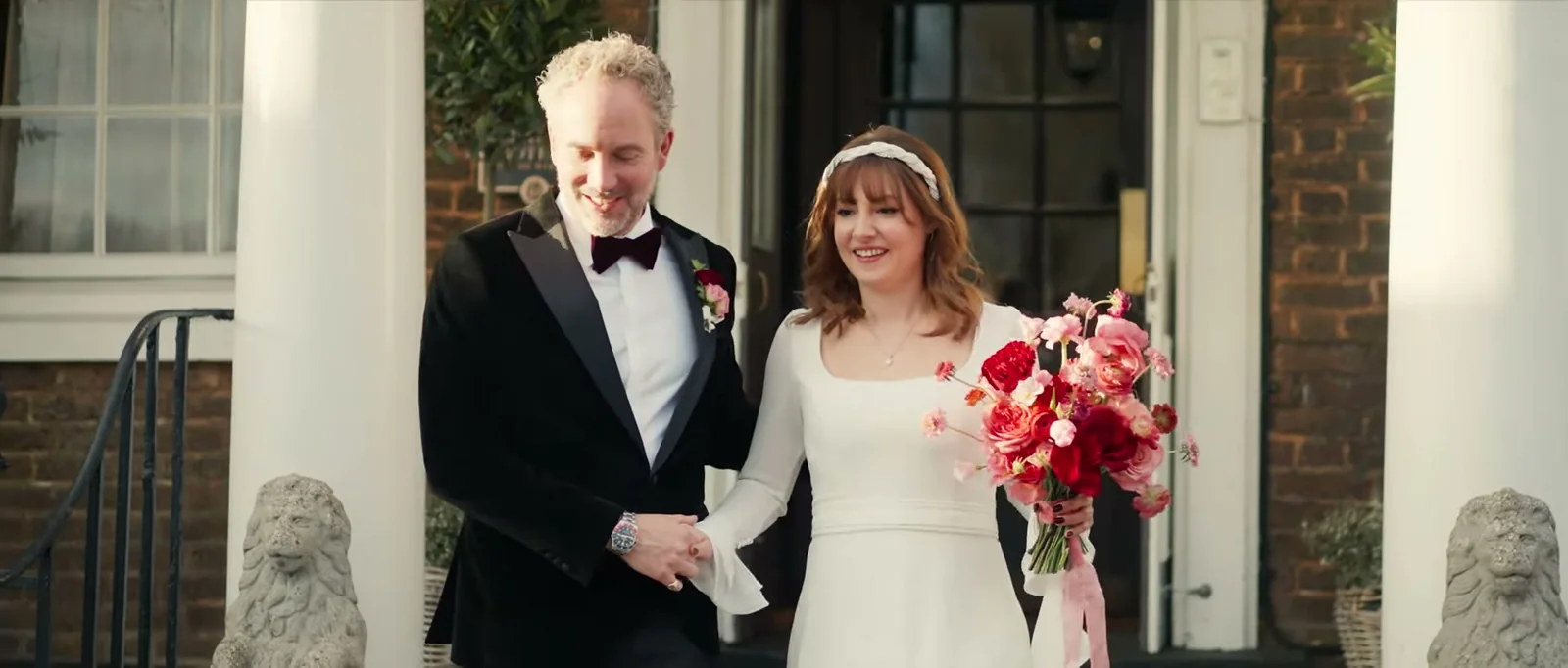 A groom and a bride with a pale headband and a bouquet walk down hand in hand between white columns, with a stone lion on each side of the steps