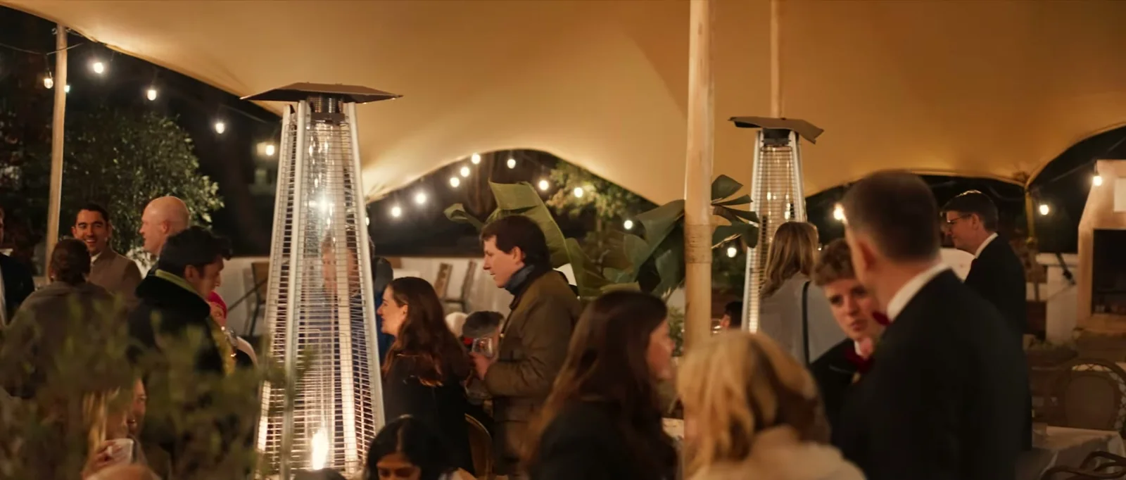 Guests in coats talk beneath a cream canopy hung with strings of lights, with tall patio heaters among them, at night