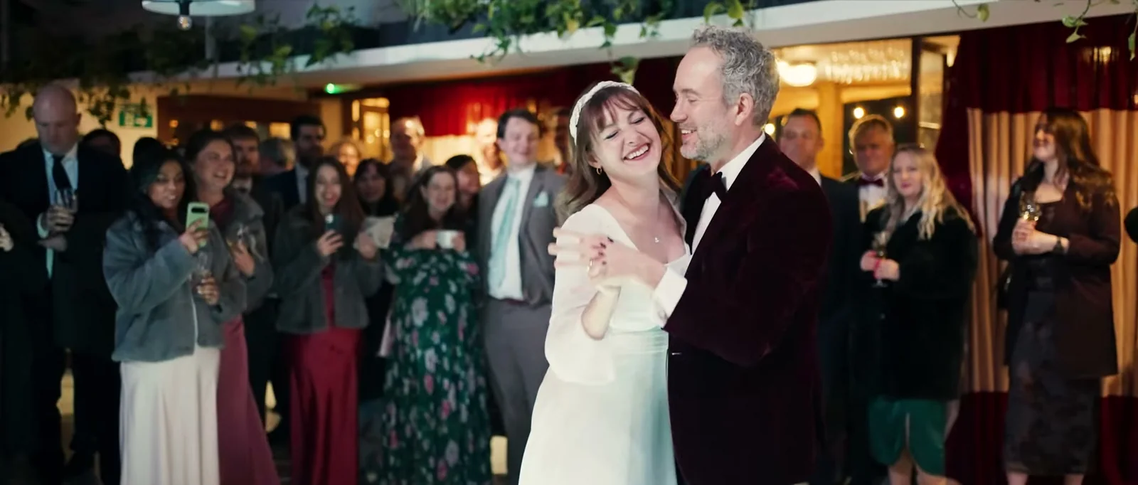 A bride laughs with her eyes closed as she dances with a groom in a burgundy velvet jacket while guests watch with phones raised