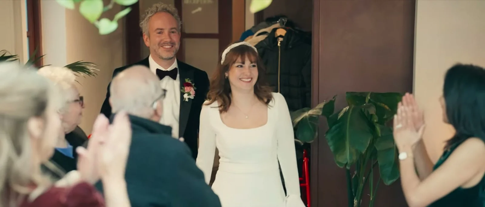 A groom and a bride with a pale headband smile as guests applaud them through a doorway