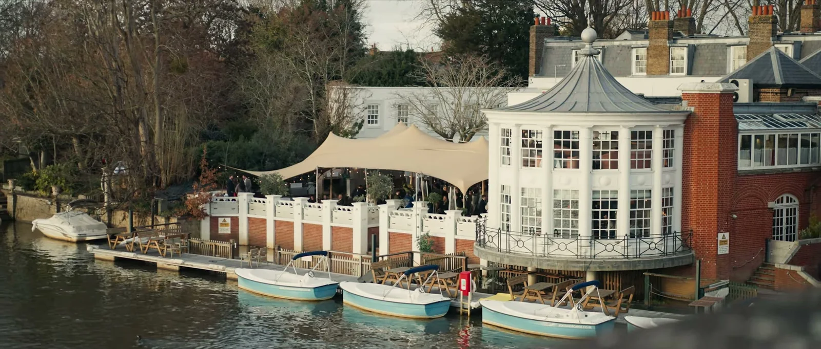 A riverside building with a round white bay, a cream canopy over a terrace and small boats moored on the Thames below