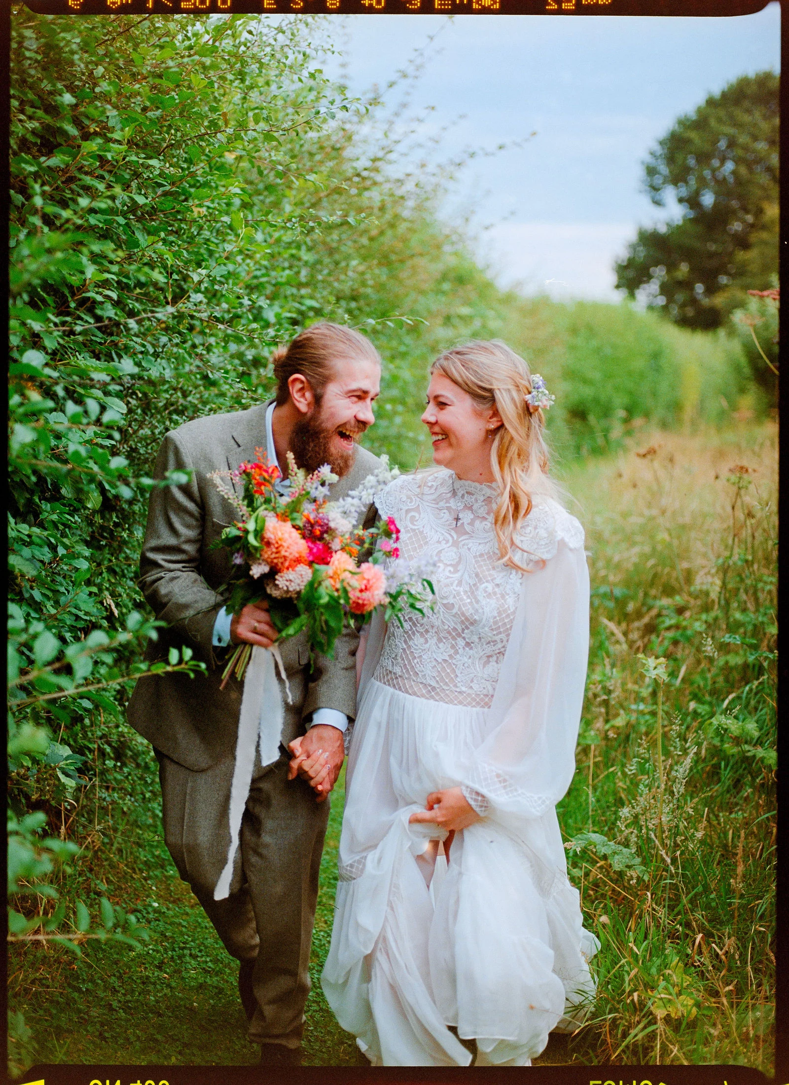 Authentic 35mm analog film photograph of Phoebe and Jamie laughing as they stroll through a wild Surrey meadow, complete with natural grain and film rebate borders