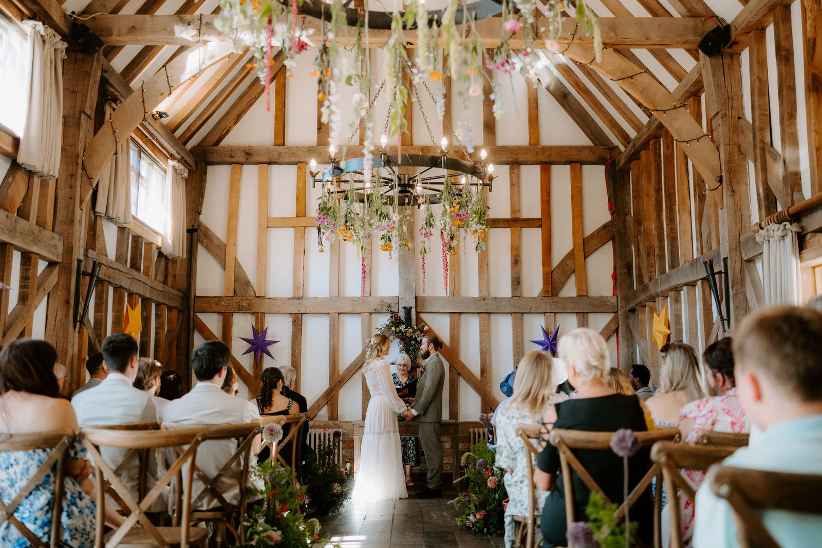 Phoebe and Jamie exchanging wedding vows beneath a floral wagon wheel chandelier inside the vaulted oak barn at Gate Street Barn in Surrey