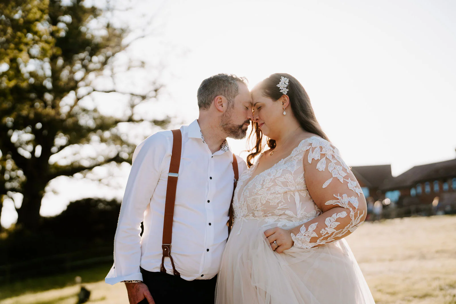 A groom and a bride in a lace gown touch foreheads with their eyes closed in low sun, with a tree on the left and a low brick building on the right at Lythe Hill in Haslemere