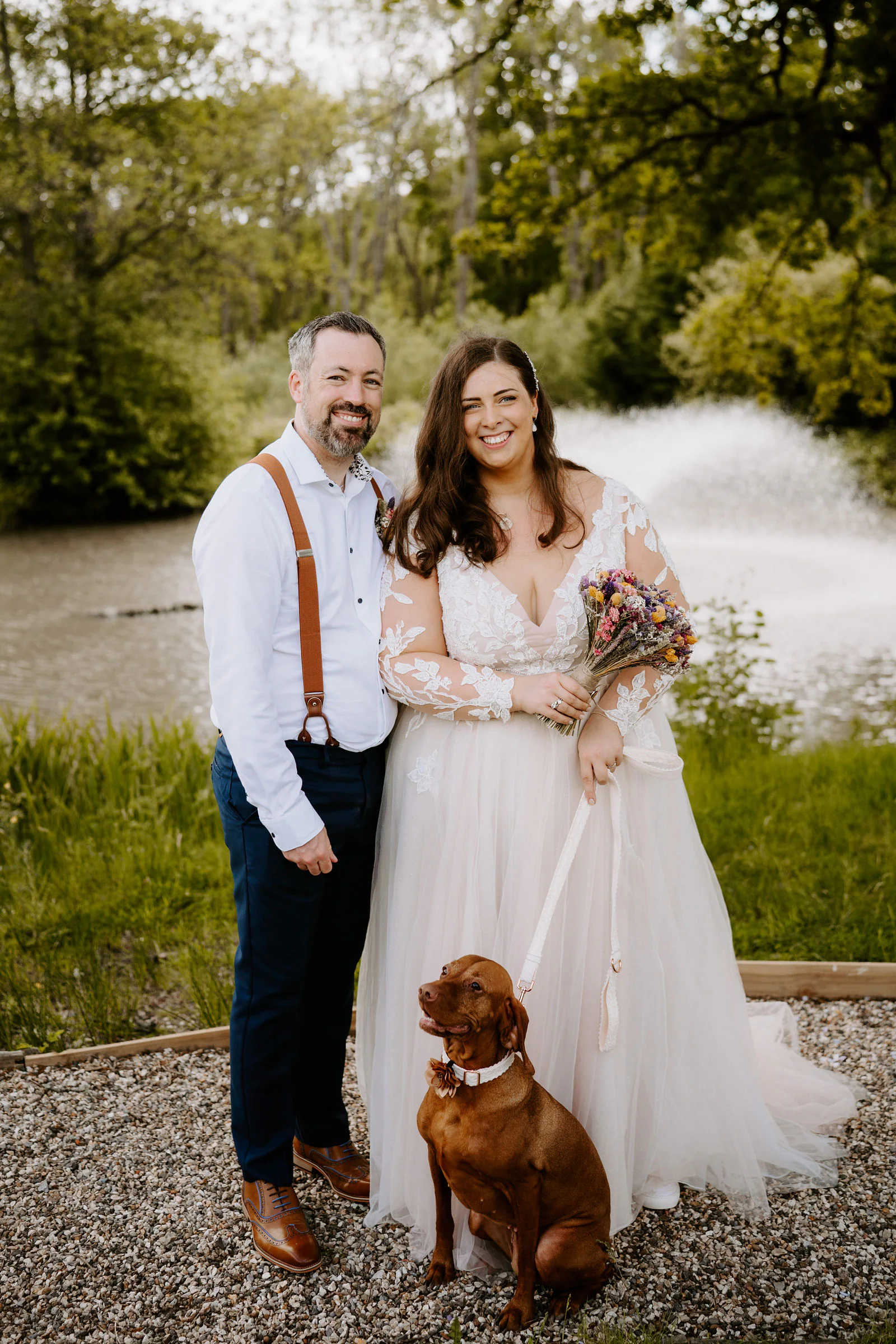 A groom in a white shirt and tan braces stands beside a smiling bride holding a bouquet of dried flowers, with a brown dog in a flower collar sitting at her feet and a fountain behind them on the lake at Lythe Hill Hotel in Haslemere
