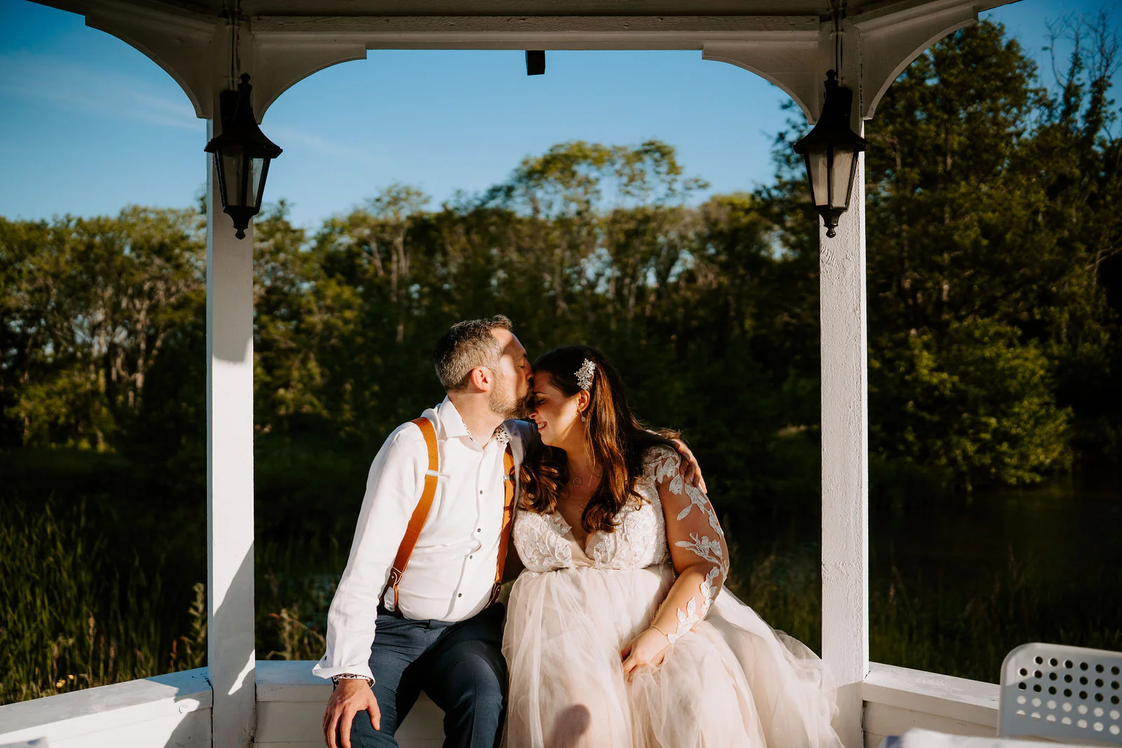 A groom kisses his bride's forehead as they sit in a white gazebo with lanterns on its posts, with trees and a lake behind them at Lythe Hill Hotel in Haslemere