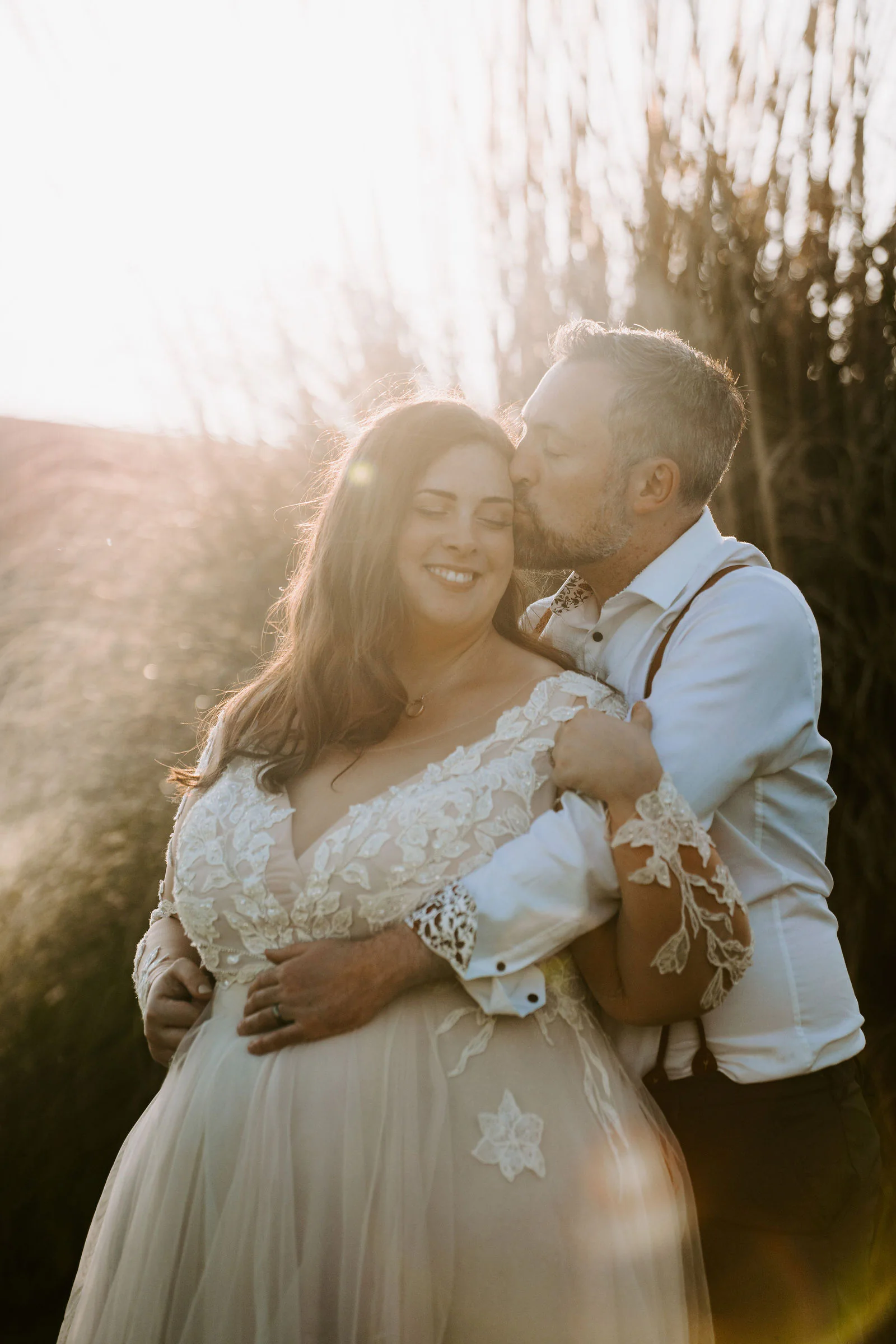 A groom holds his smiling bride close and kisses her cheek as the sun shines through tall grasses behind them at Lythe Hill in Haslemere