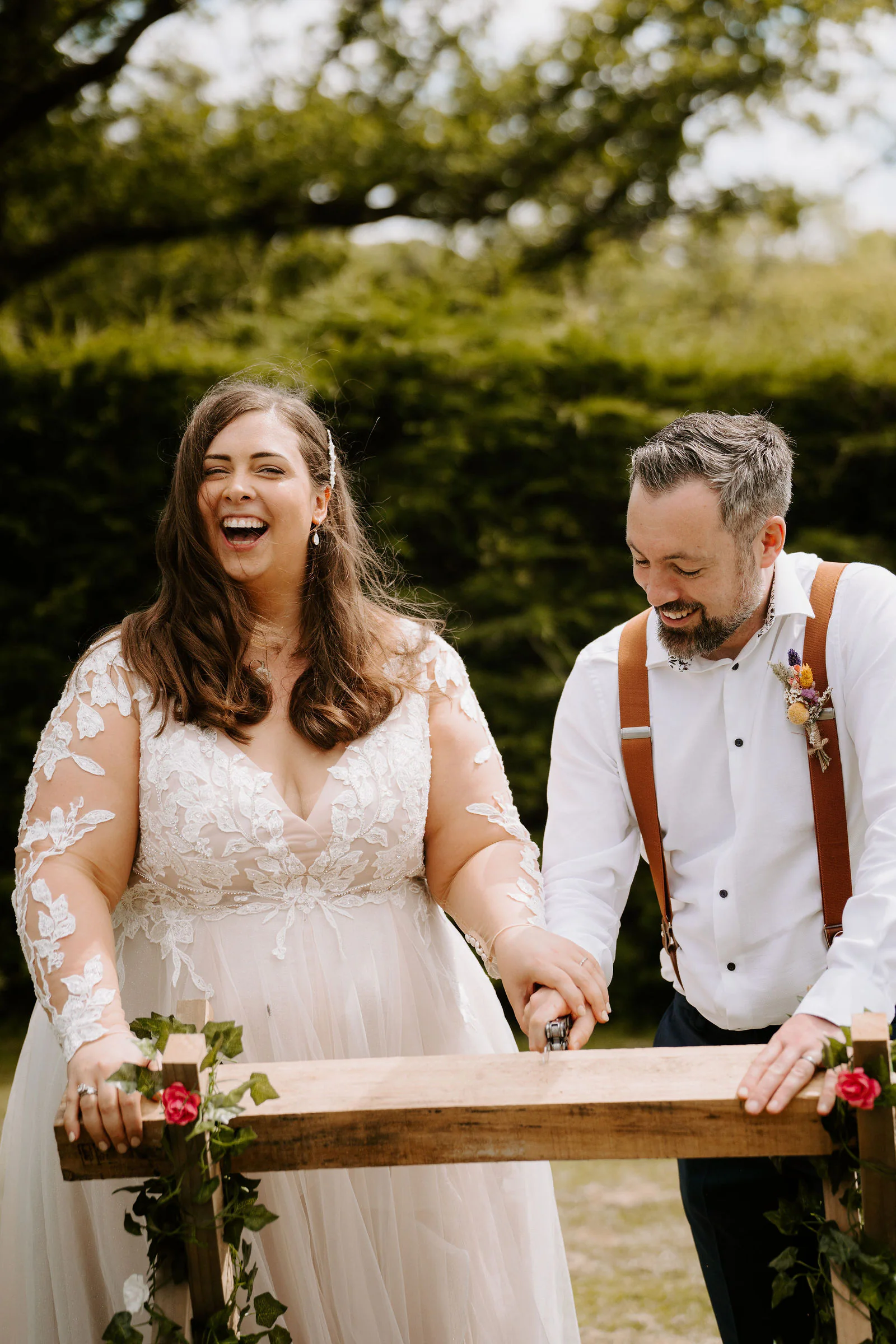 A bride in a lace gown laughs aloud as she and her groom saw through a log on a wooden trestle trimmed with ivy and red roses, with a hedge behind them at Lythe Hill in Haslemere