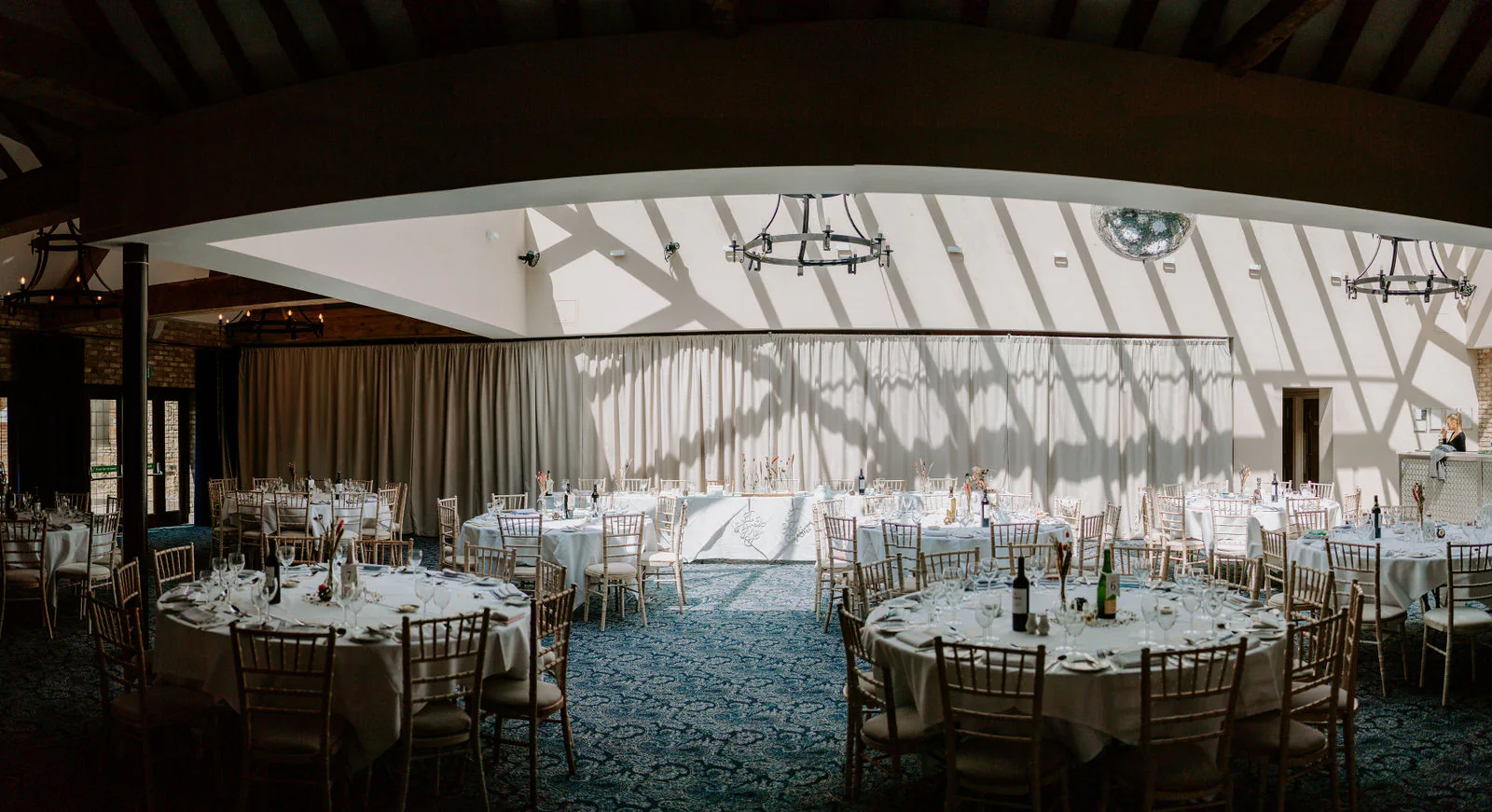 Round tables laid with glasses and wooden chairs in a wedding breakfast room with sunlight and shadows across a long curtain, a mirror ball and iron chandeliers at Lythe Hill Hotel in Haslemere