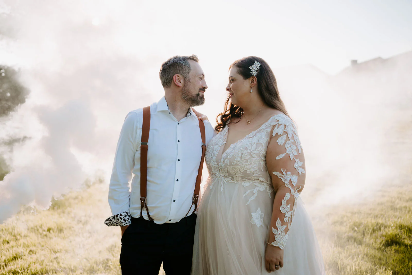 A groom in a white shirt and tan braces and a bride in a lace gown look at each other while white smoke drifts across the grass behind them at Lythe Hill in Haslemere