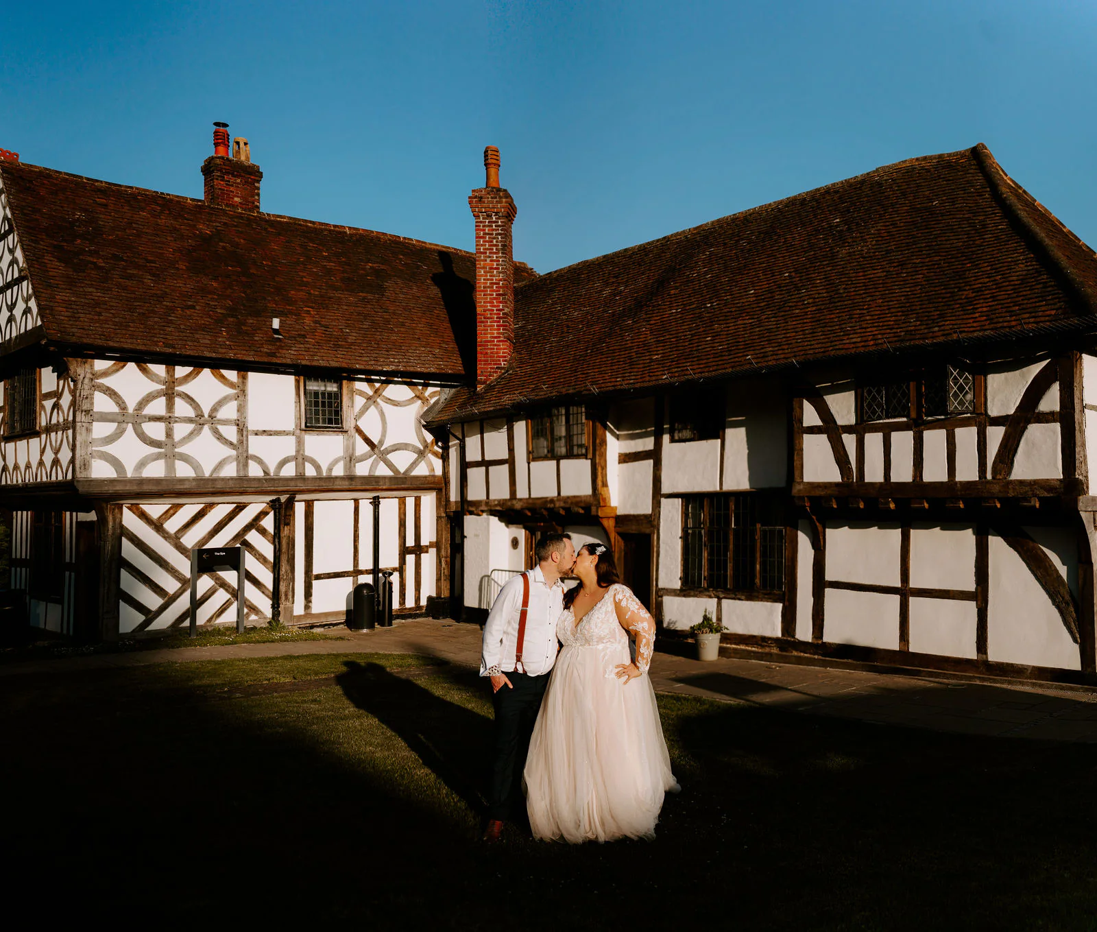 A bride and groom kiss on a lawn in front of a black and white timber-framed building with a tiled roof and brick chimneys under a deep blue sky at Lythe Hill Hotel in Haslemere