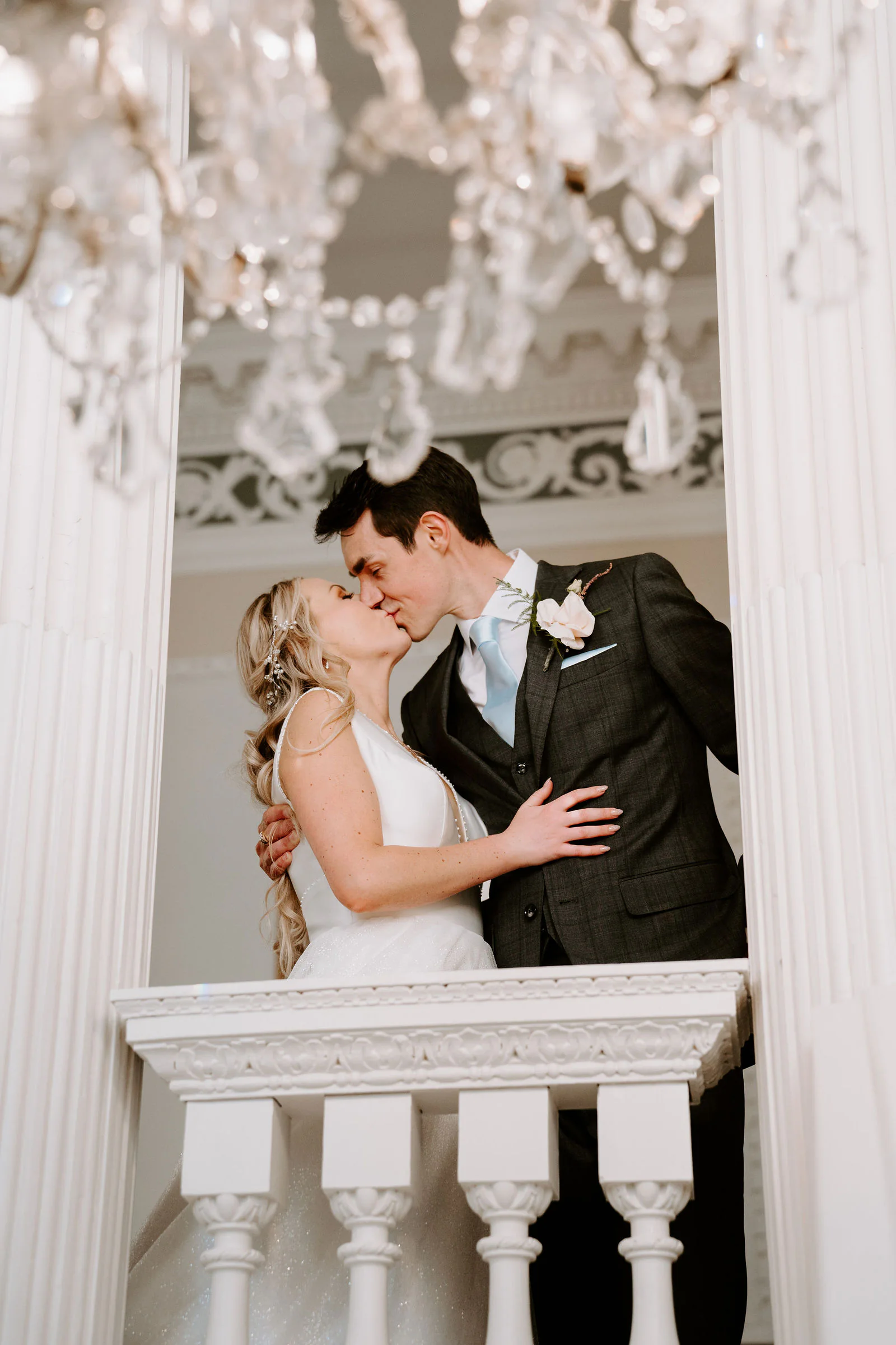A groom and a bride kiss on a white balustraded balcony between pillars beneath crystal chandeliers at Buxted Park Hotel