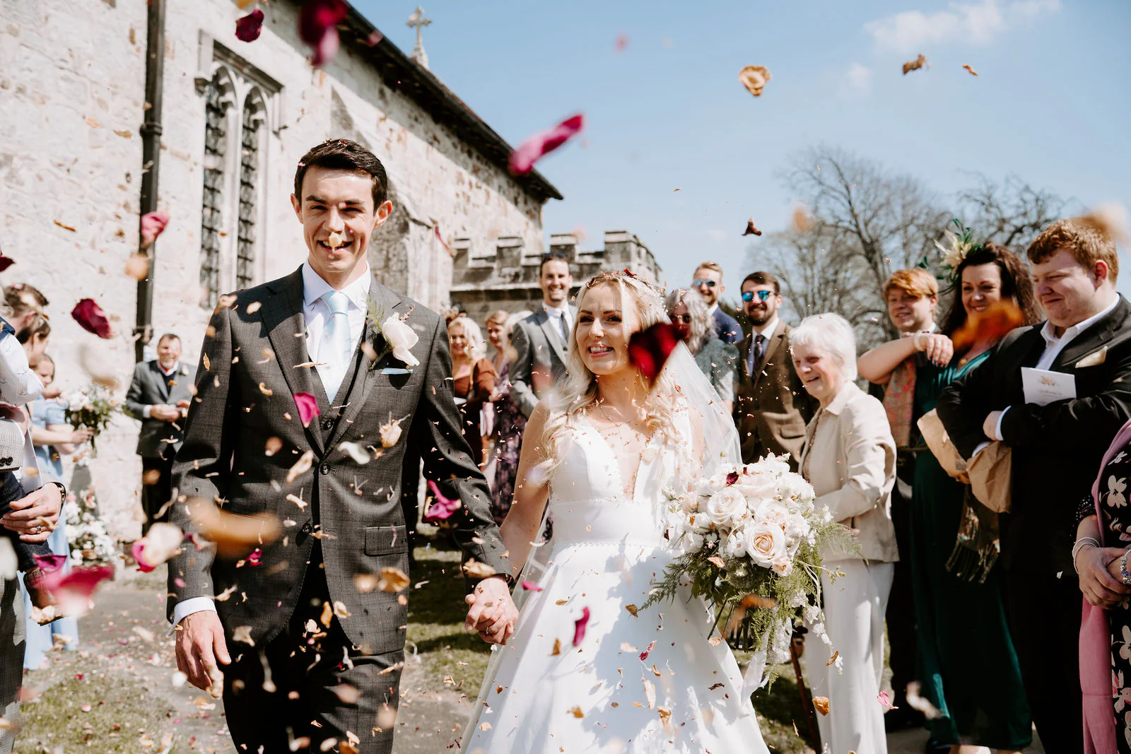 A bride and groom walk hand in hand through a shower of petals beside a stone church as guests smile behind them after their Buxted Park Hotel wedding ceremony