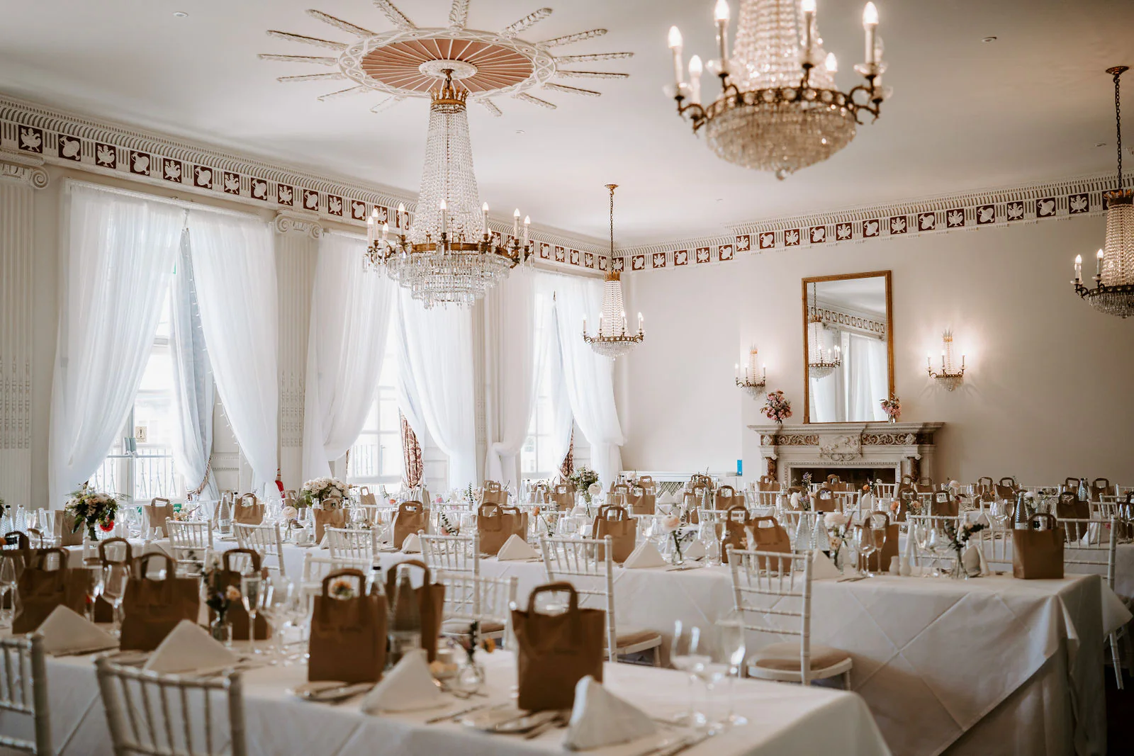 A wedding breakfast room with long white-clothed tables, white chairs, crystal chandeliers and tall windows with white curtains at Buxted Park Hotel