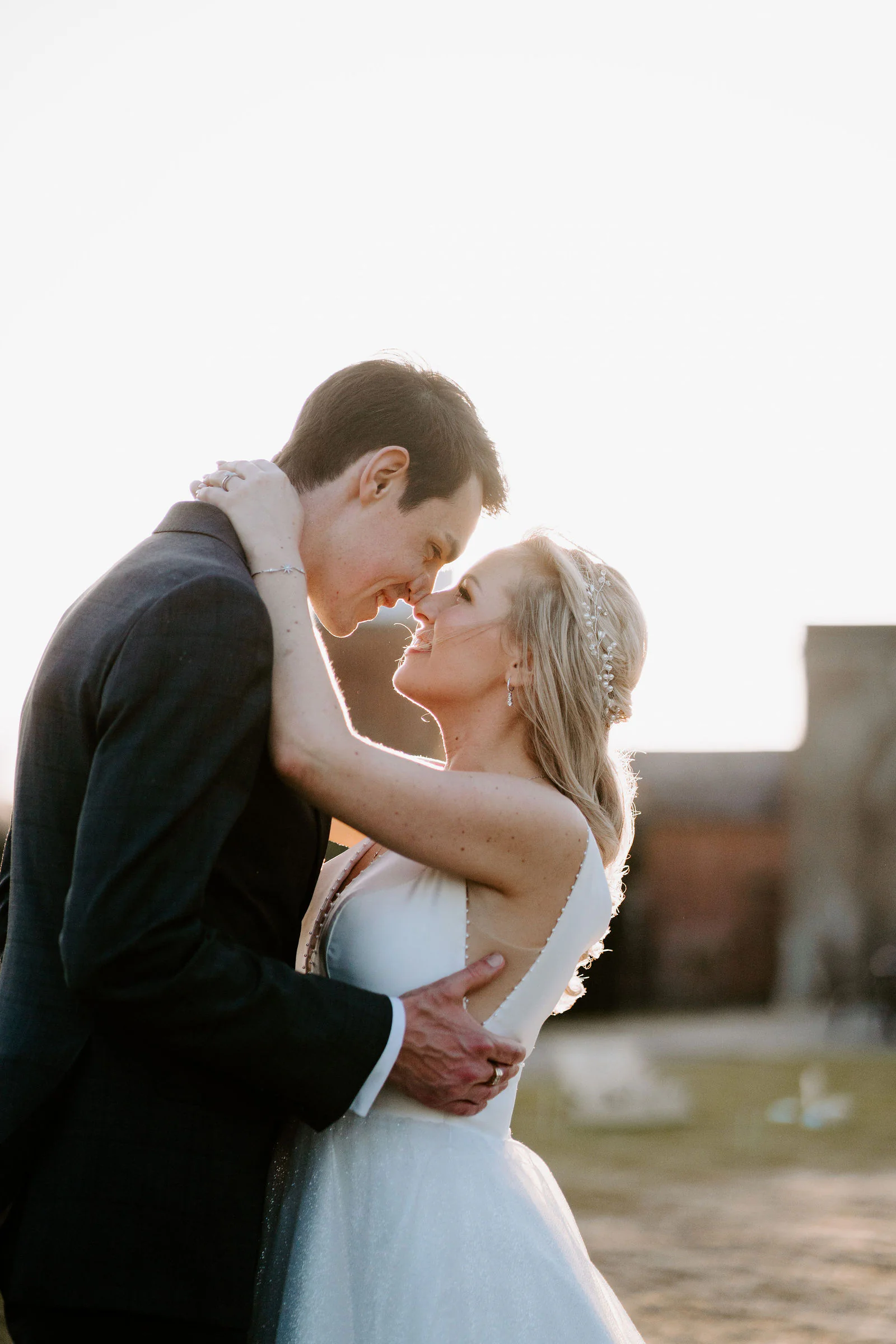 A bride and groom touch noses in low backlit sunlight with a softly blurred old building behind them at Buxted Park Hotel