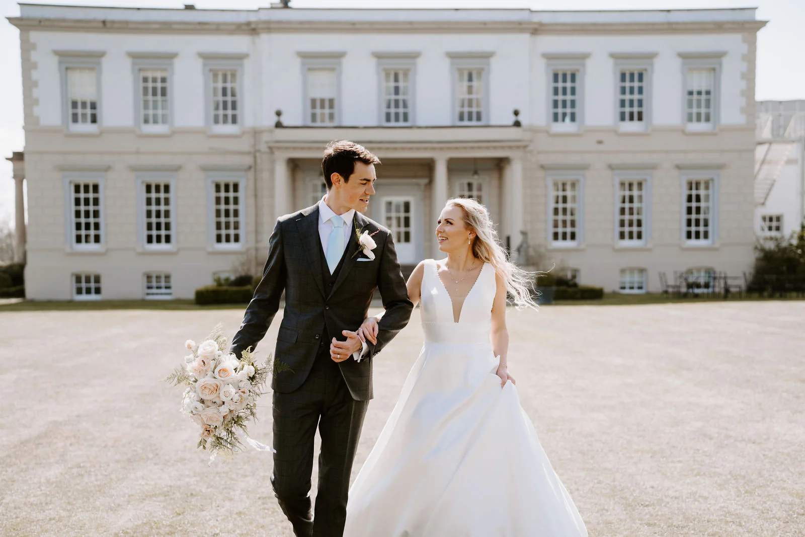 A groom in a grey check suit and a bride in a white gown walk arm in arm across a gravel lawn, looking at each other, with the pale front of Buxted Park Hotel behind them